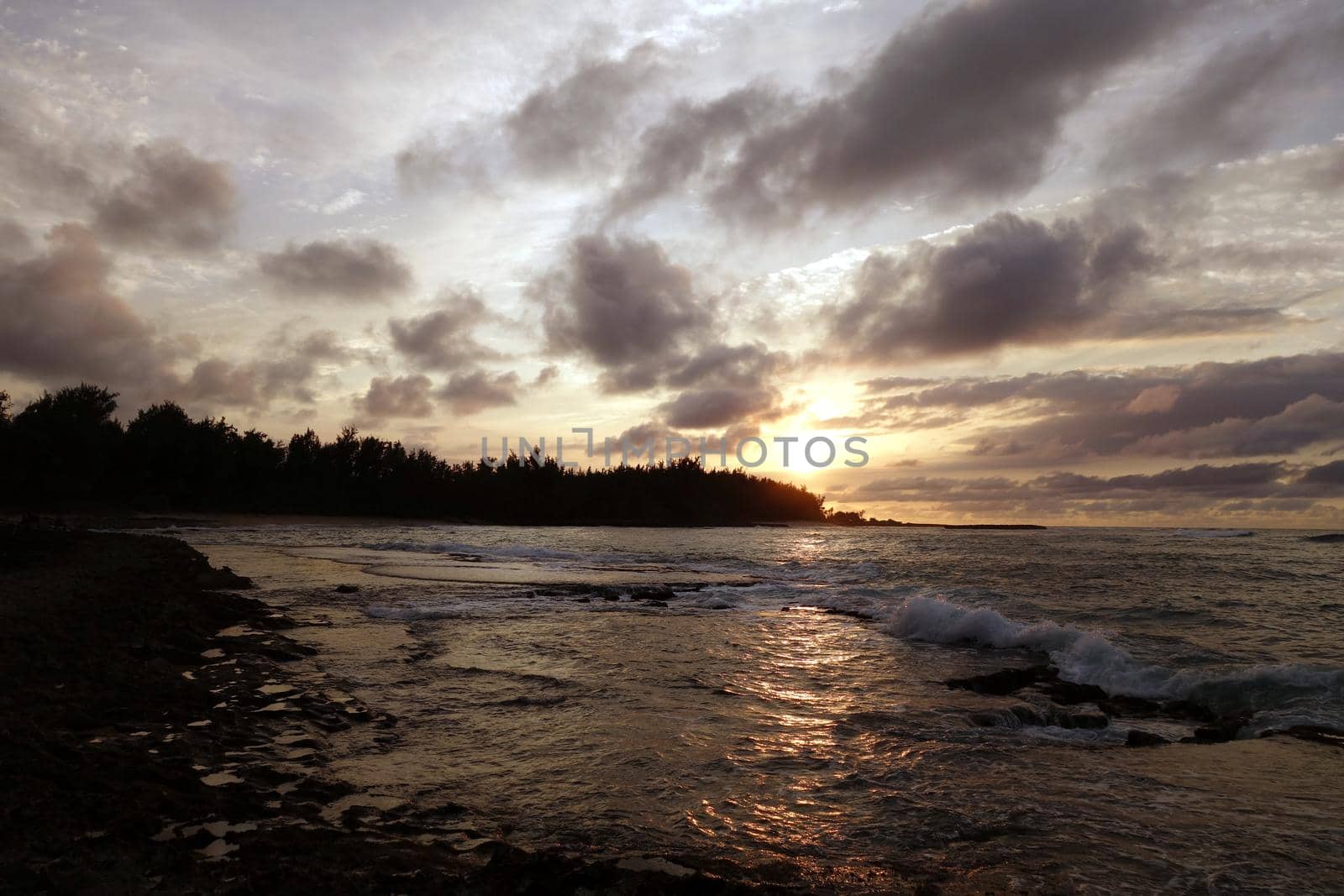 Sunset through the clouds and over the trees as it reflects on the watery waves at Kawela Bay rocky shore by EricGBVD