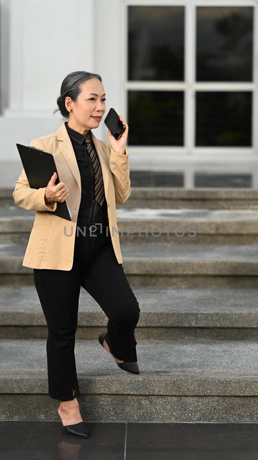 Full length portrait of mature businesswoman talking on cell phone and standing in front of an office building.