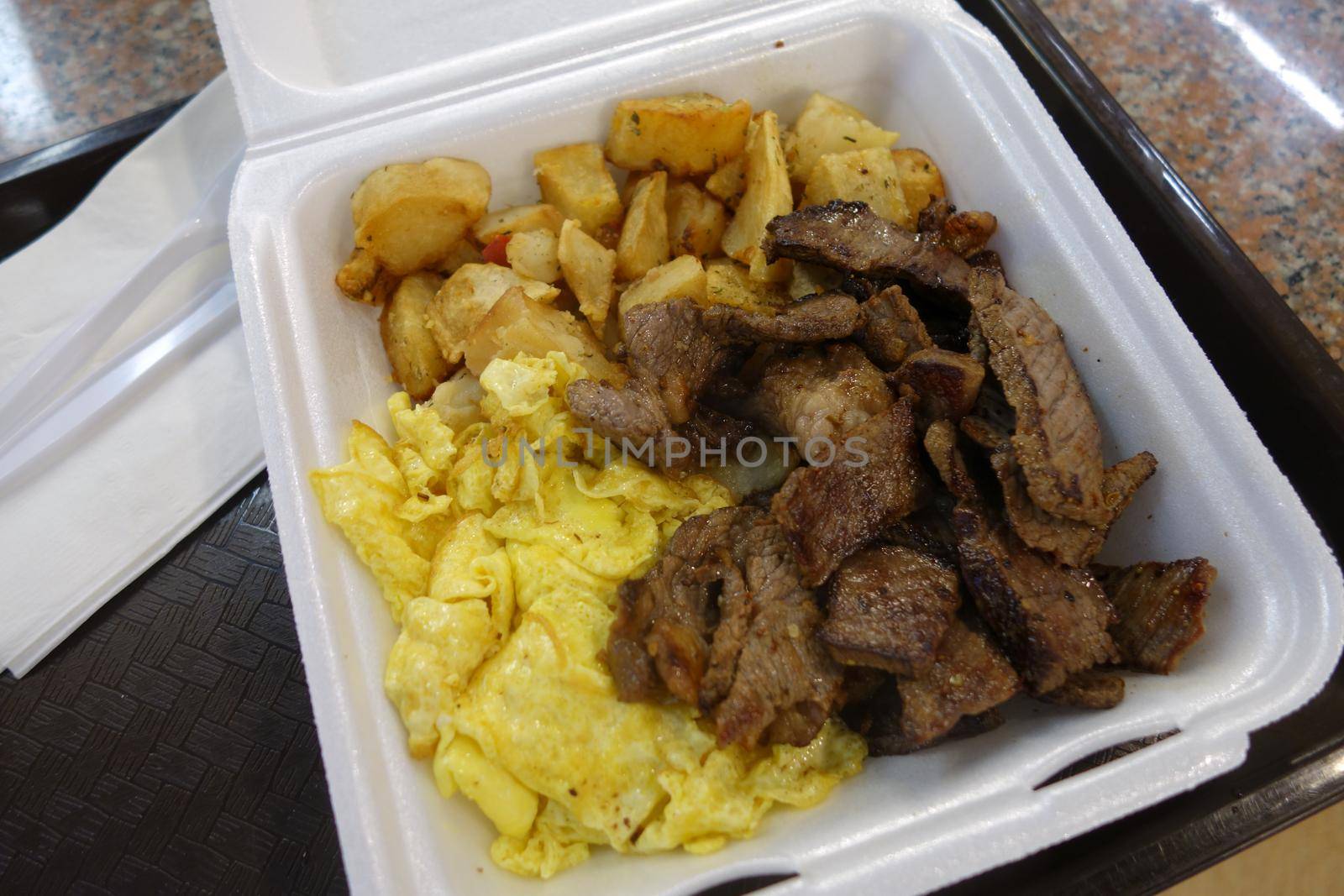 Kiawe grilled steak, scrambled eggs, country fried potatoes in a styrofoam plate on a tray on a table.