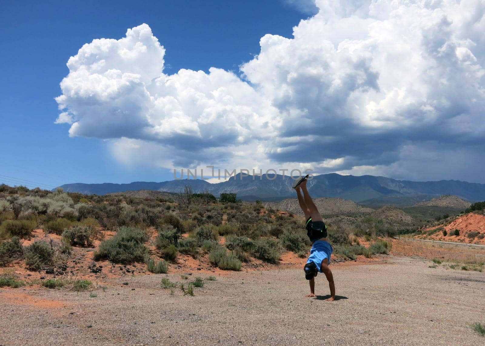 Man wearing a hat, t-shirt, shorts, and slippers does Handstands in the desert Mountain with dramatic clouds in sky in Utah, USA.