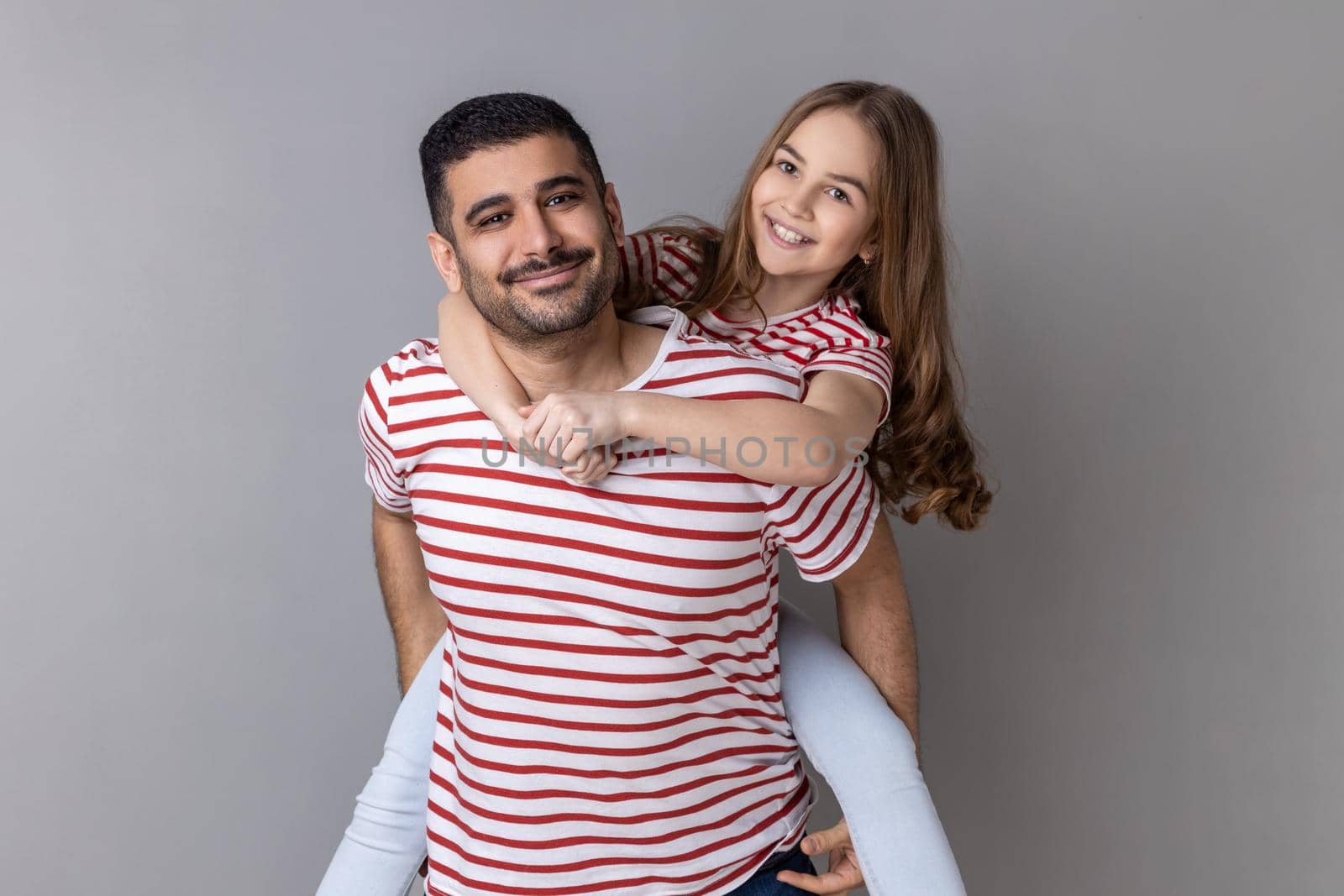 Portrait of positive optimistic father and daughter in striped T-shirts spending time together, dad holding his charming little kid on his back. Indoor studio shot isolated on gray background.