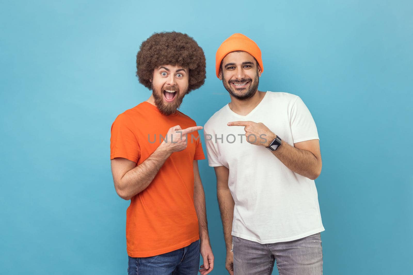Best friends. Portrait of two young adult hipster men pointing finger each other and looking at camera with positive joyful expressions. Indoor studio shot isolated on blue background.