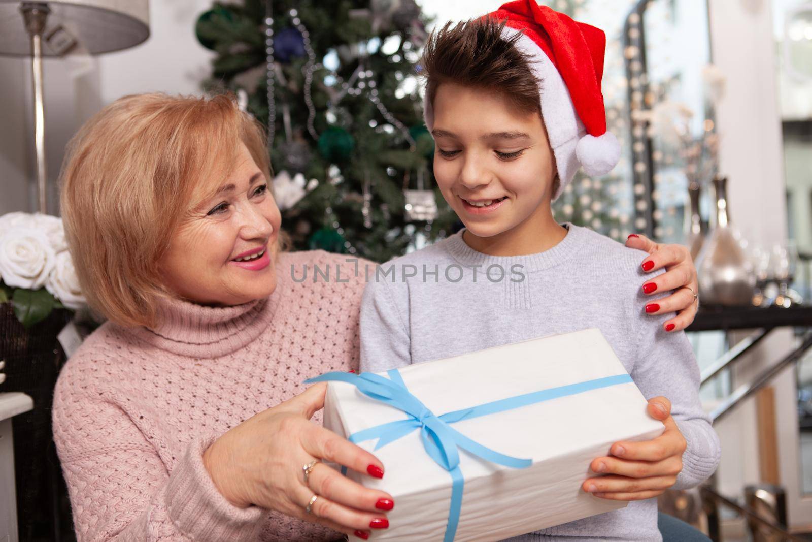 Elderly woman smiling joyfully, giving Christmas present to her young grandson. Cute little boy in Santa Claus hat receiving x-mas gift from grandma
