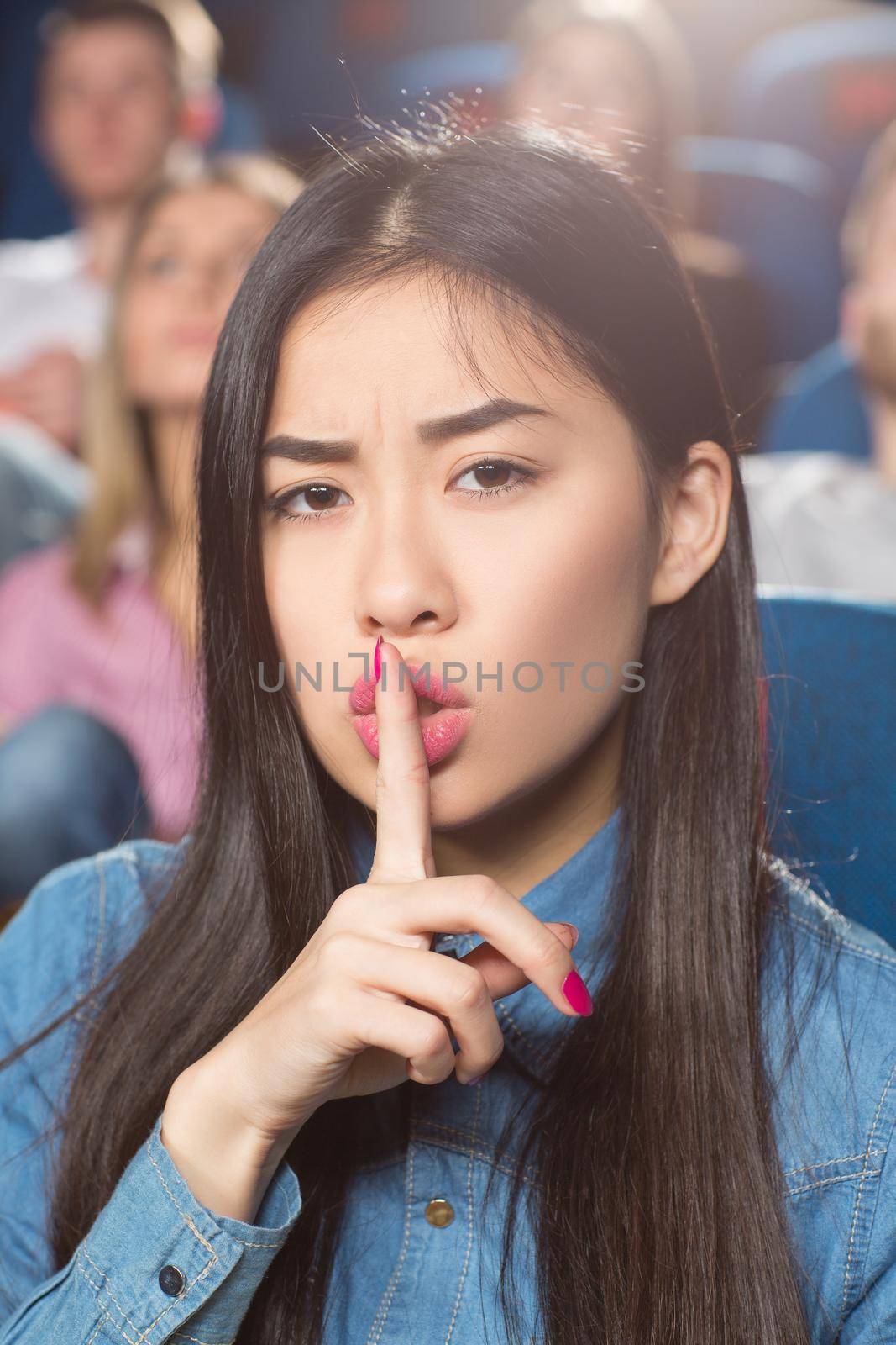 Be quiet. Gorgeous young asian woman making shushing gesture with her finger to her lips while watching movies at the cinema