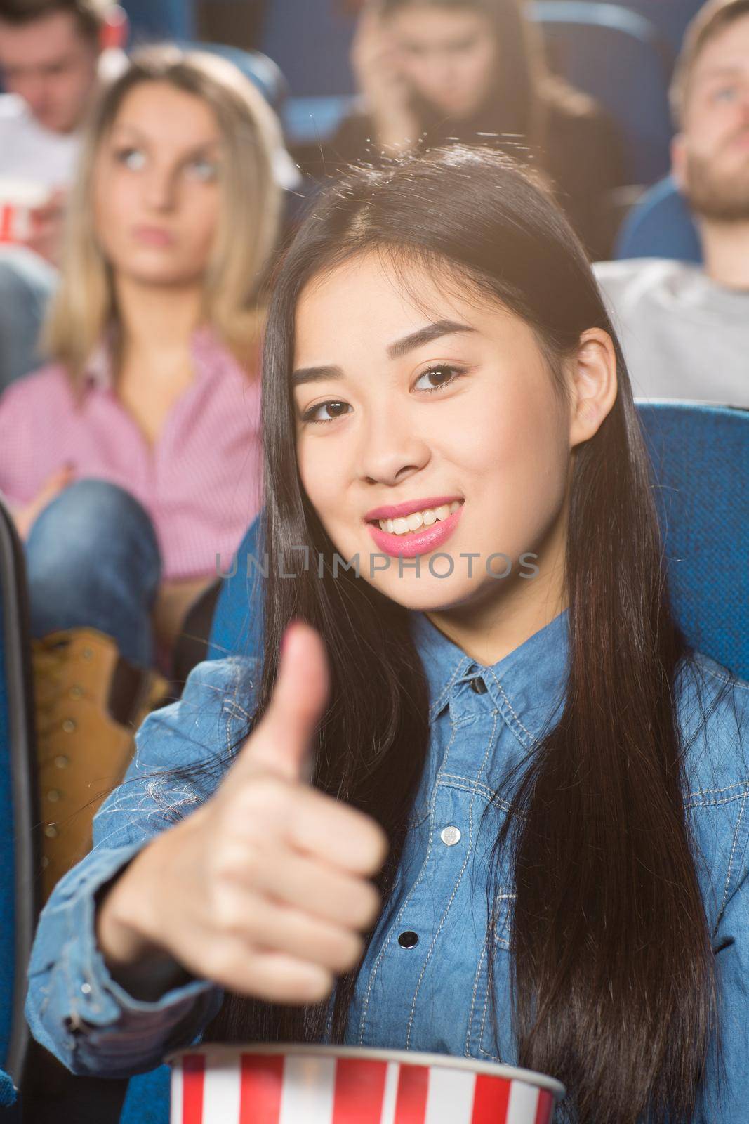 Movie approved. Young happy asian woman holding popcorn bucket showing thumbs up smiling cheerfully at the cinema