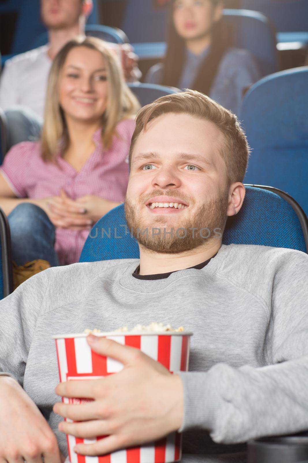 Cinema lover. Vertical shot of a handsome bearded young man holding popcorn bucket laughing watching a movie at the local cinema