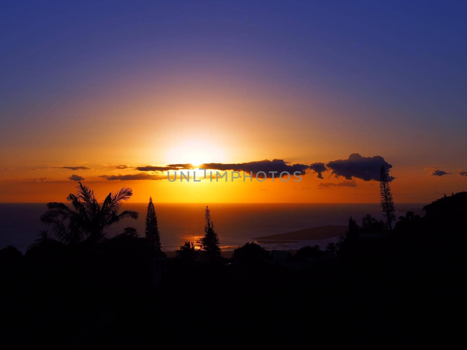 Sunset through the clouds over the pacific ocean with light reflecting on water past tropical silhouette of trees on Oahu, Hawaii.  February 2016.