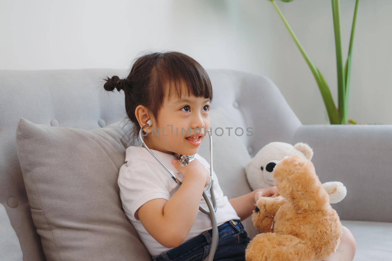 Little girl pretending to be doctor while treating her stuffed toy with stethoscope on sofa in living room at home by makidotvn