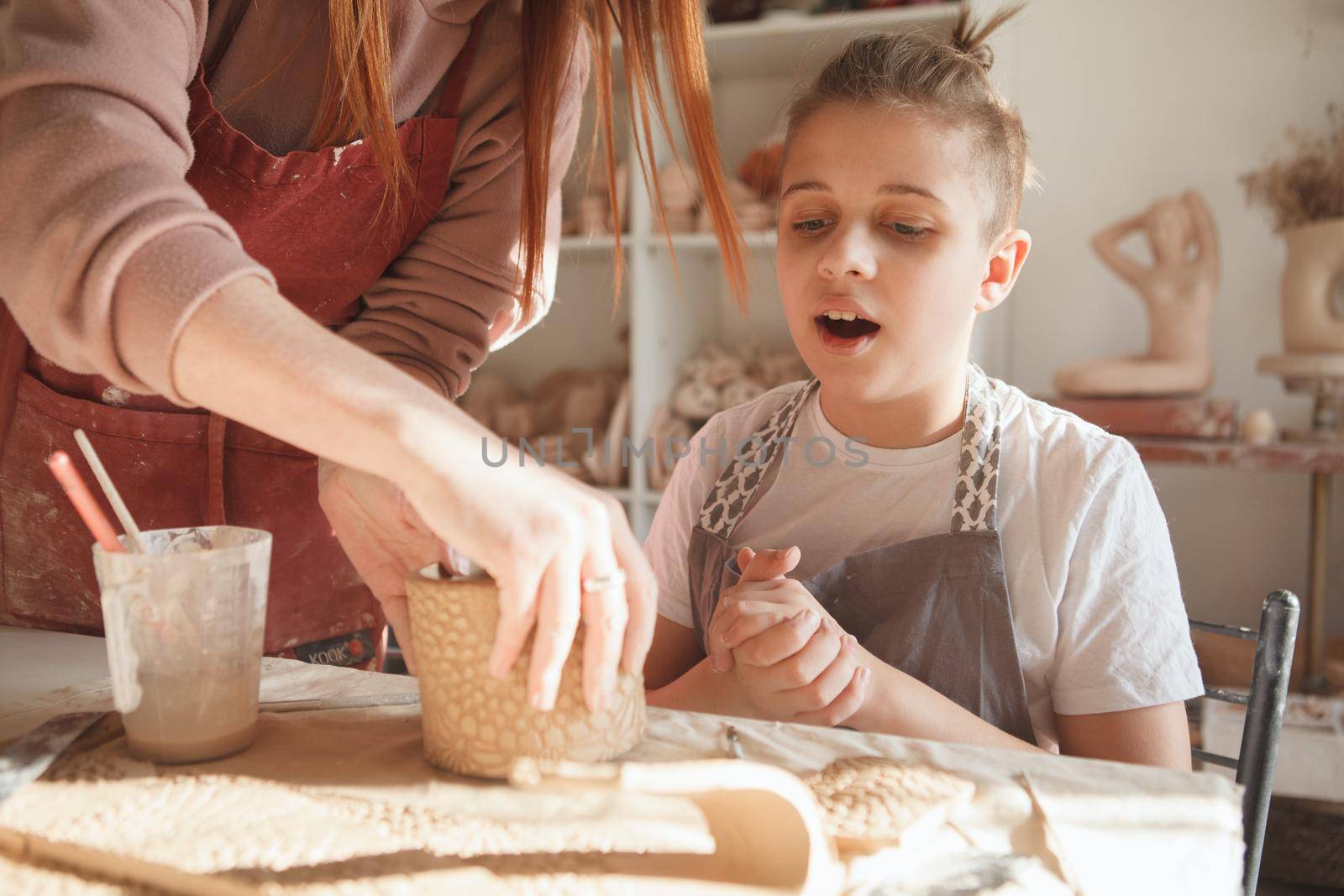 Young boy looking excited, watching professional ceramist helping him making tea mug at pottery class