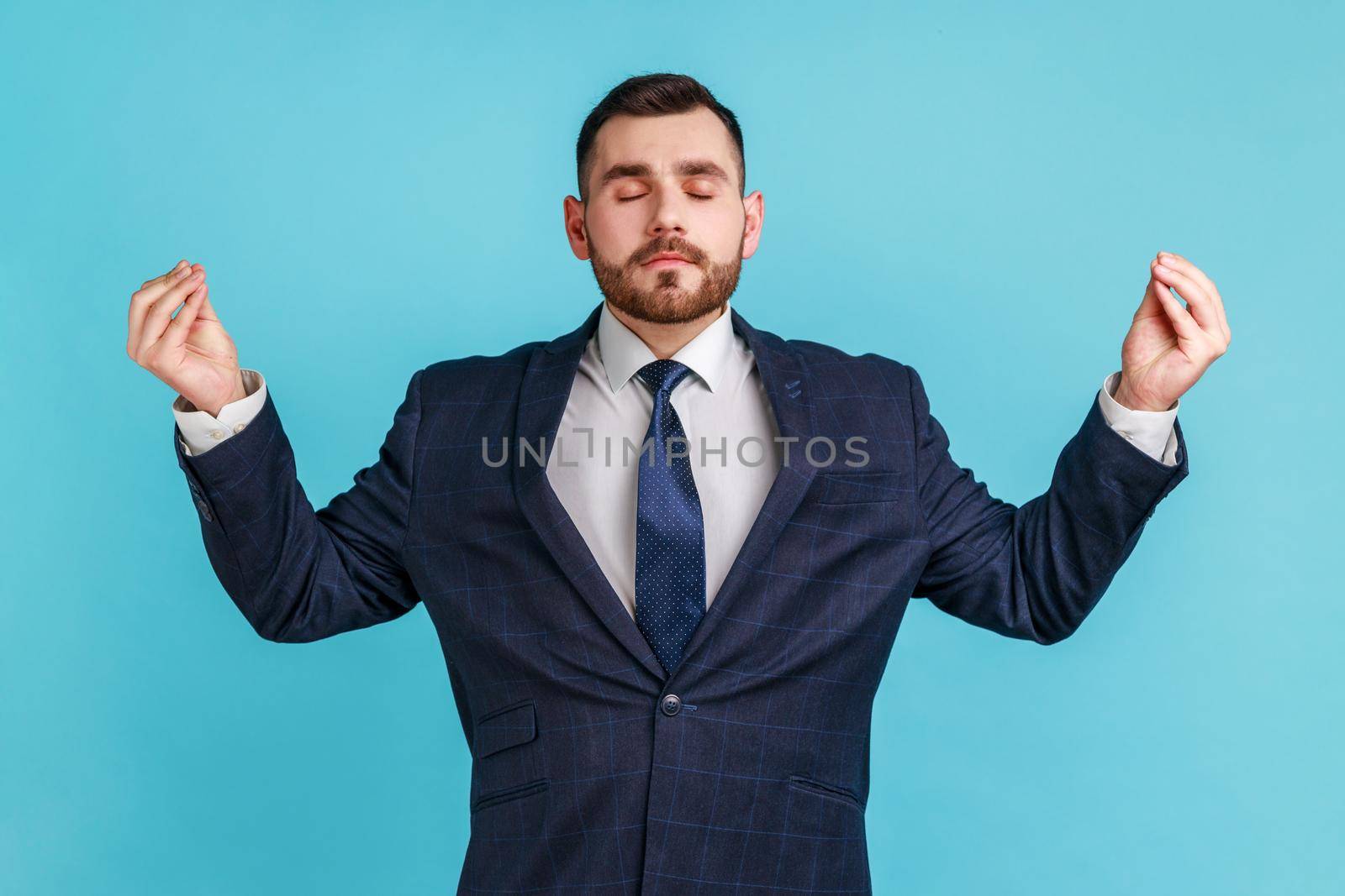 Portrait of calm relaxed handsome bearded young adult man wearing official style suit raised arms and doing yoga meditating exercise. Indoor studio shot isolated on blue background.