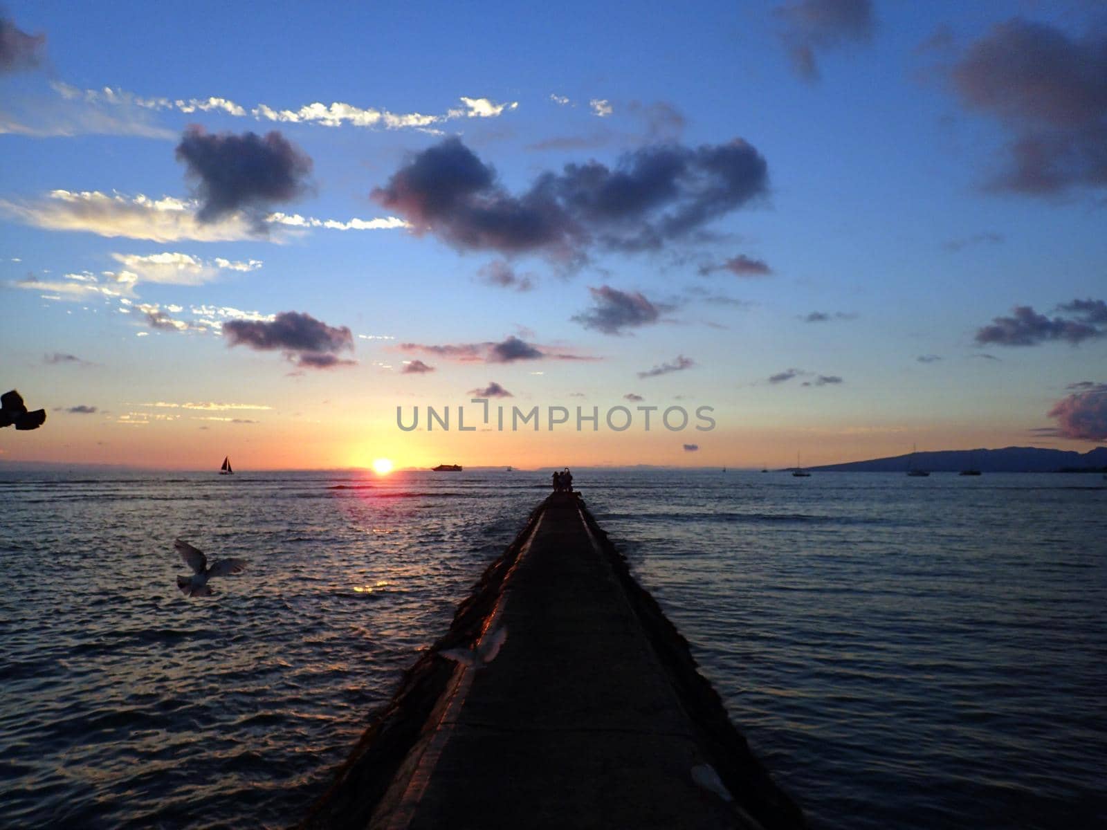 Rock Pier leads to Sunset over the Pacific ocean on the water with birds flying in the foreground and boats off the coast of Oahu, Hawaii with Waianae Mountain range visible. February 2016.