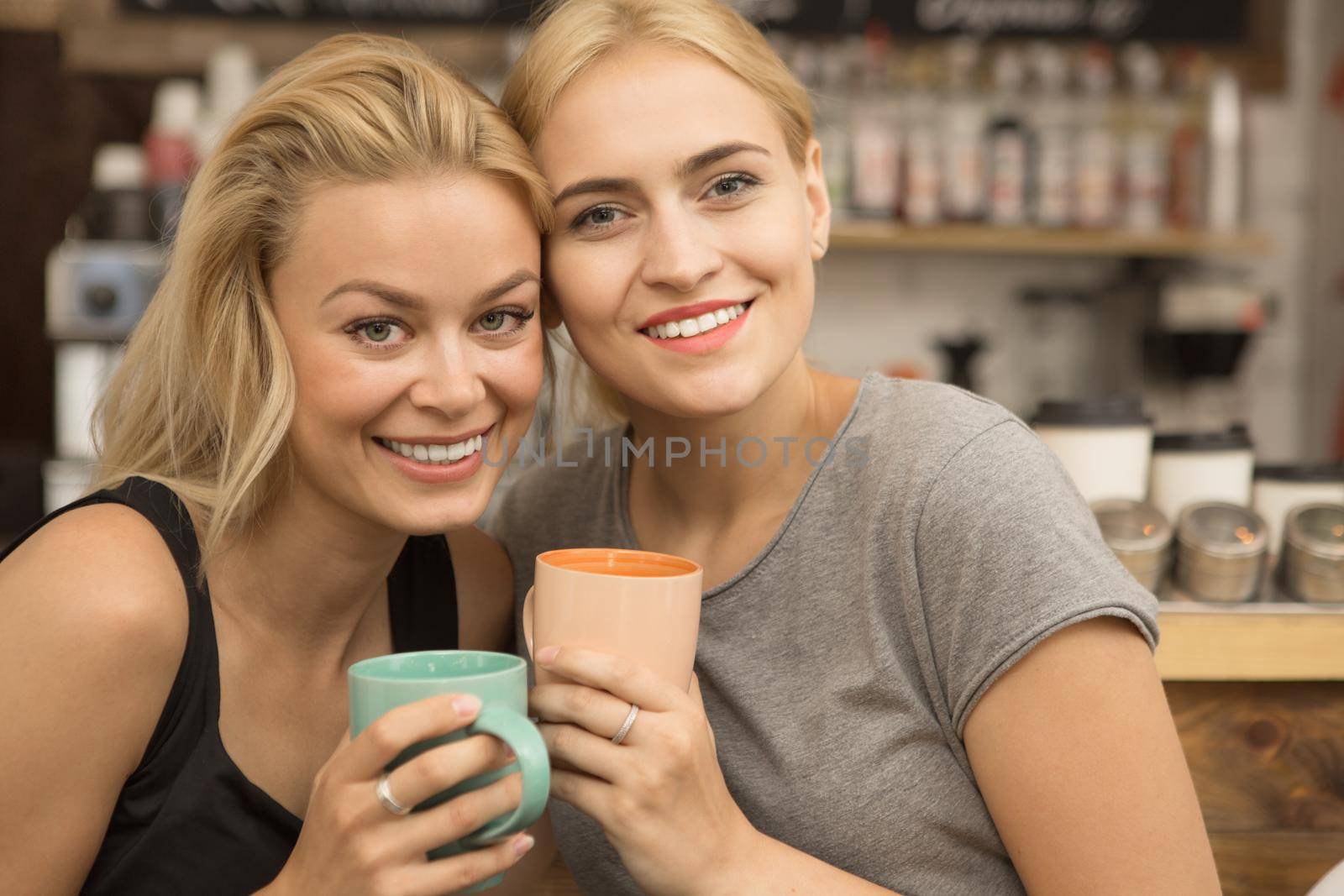 Gorgeous ypung girls smiling to the camera holding cups of coffee while relaxing together at the cafe friendship relationships students campus college women beauty expressive emotions people lifestyle weekend