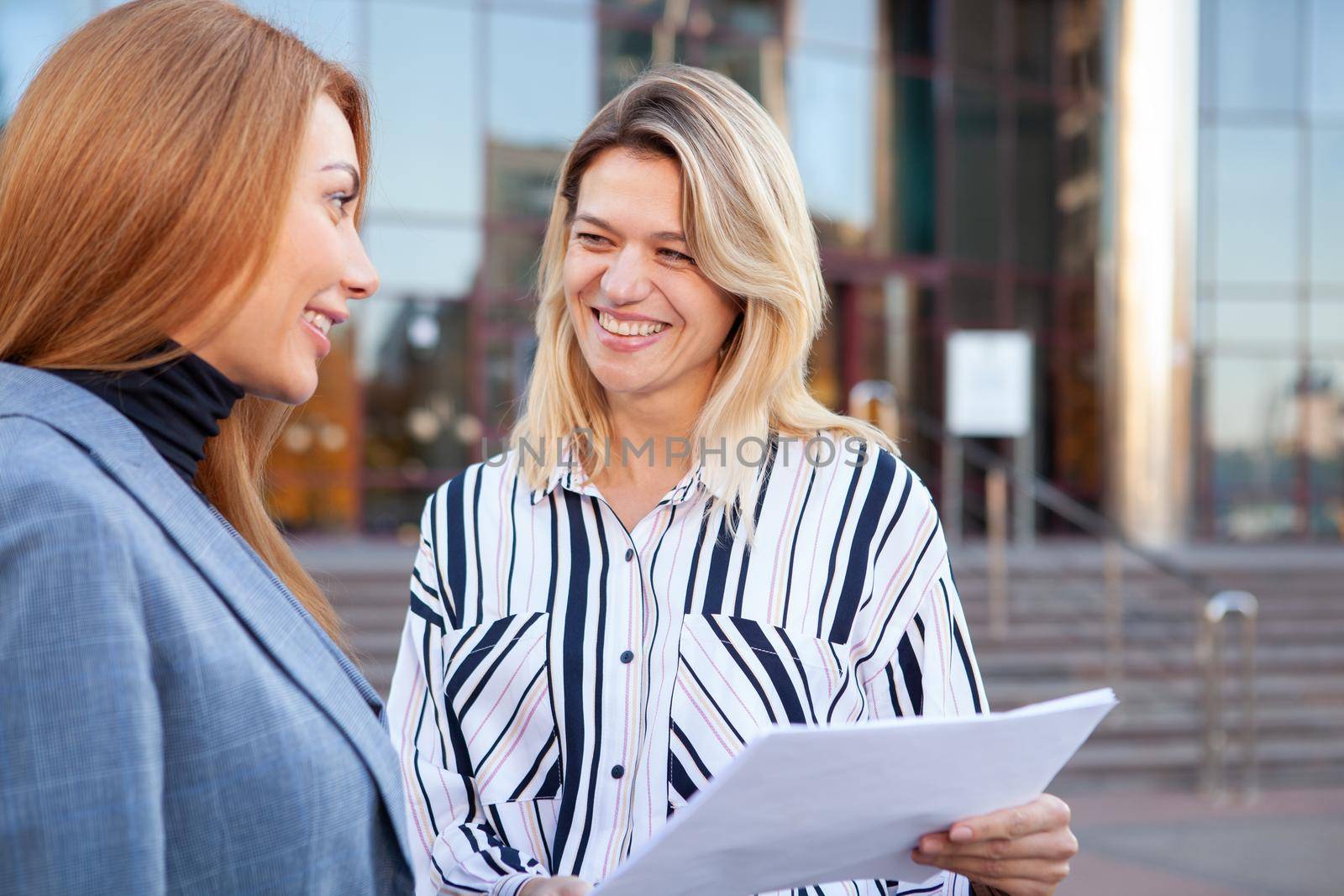 Charming mature businesswoman discussing startup with her colleague. Female entrepreneurs talking outdoors in front of office building