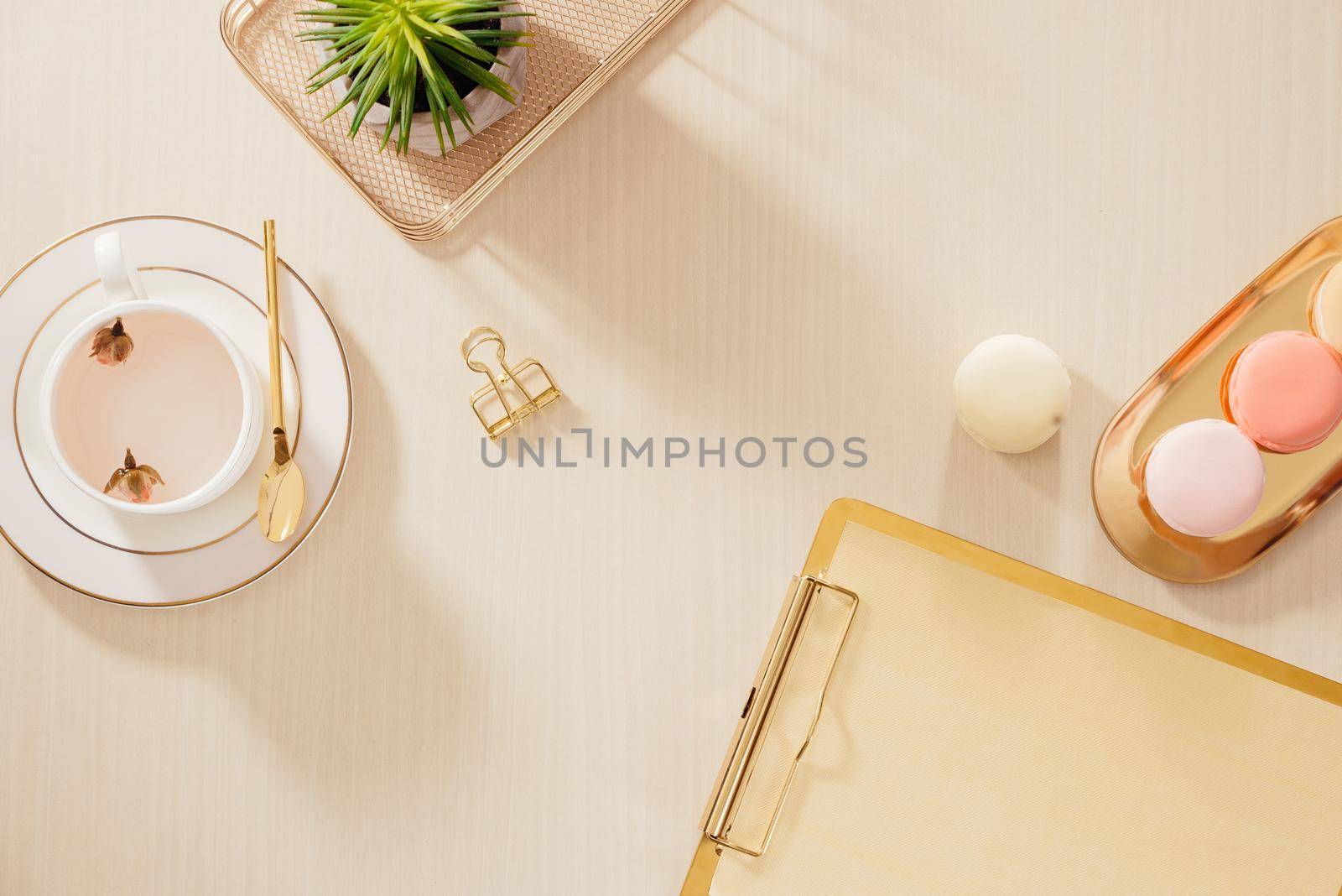 Modern gold stylized home office desk with folder, macaroons, coffee mug on beige background. Flat lay, top view lifestyle concept.`