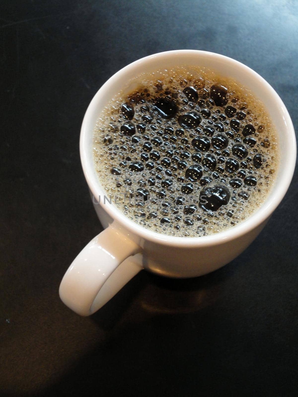 White Mug Full of Bubbly Coffee on a reflective table.                               