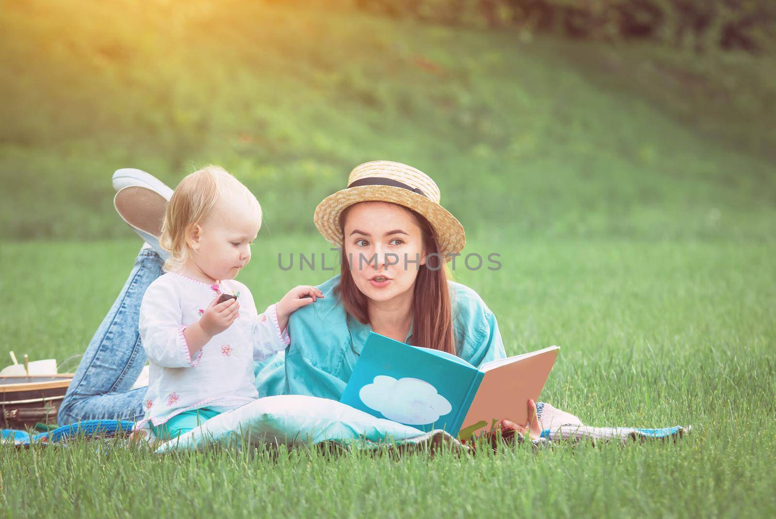 Mother is reading book for baby girl lying on the grass in the park by maramorosz