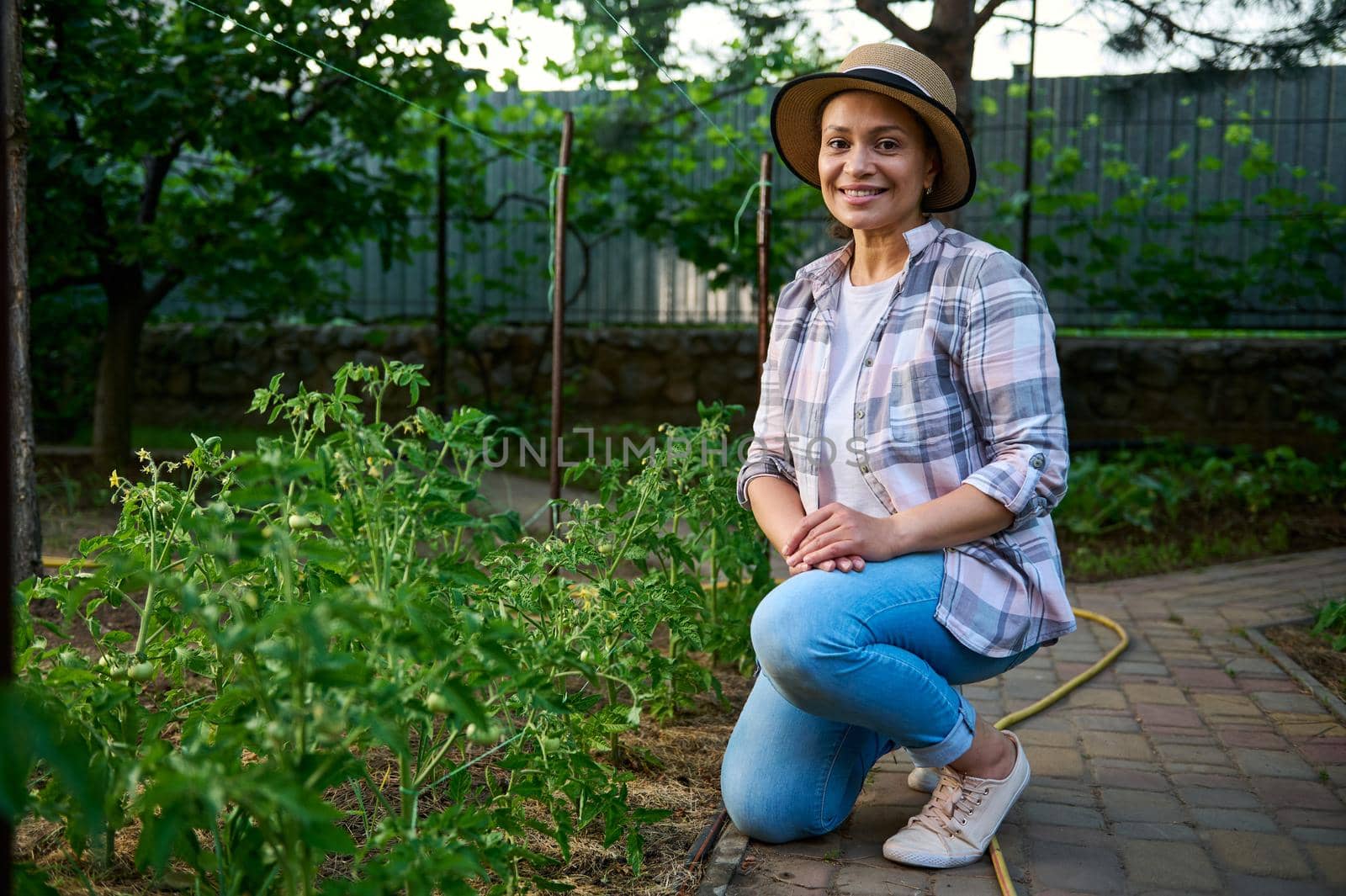Attractive successful woman, owner of an ecological farm, sits next to tomato seedlings and smiles sweetly, looking at the camera. Beautiful prosperous young female farmer enjoying gardening in spring