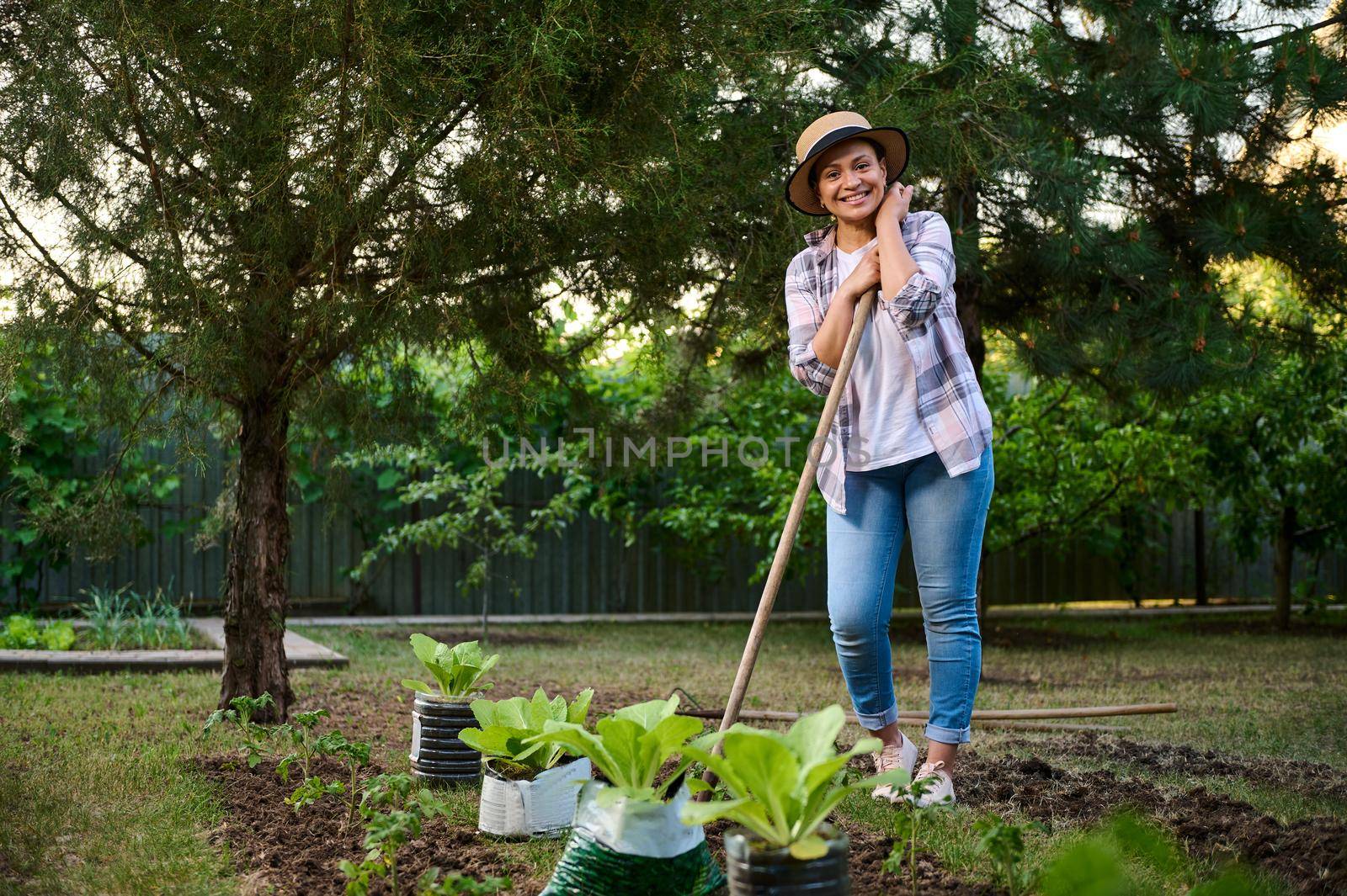 Charming multi-ethnic woman, gardener smiles at the camera, posing with a hoe while gardening in the ecological vegetable garden in the countryside house. Portrait of an inspired farmer horticulturist