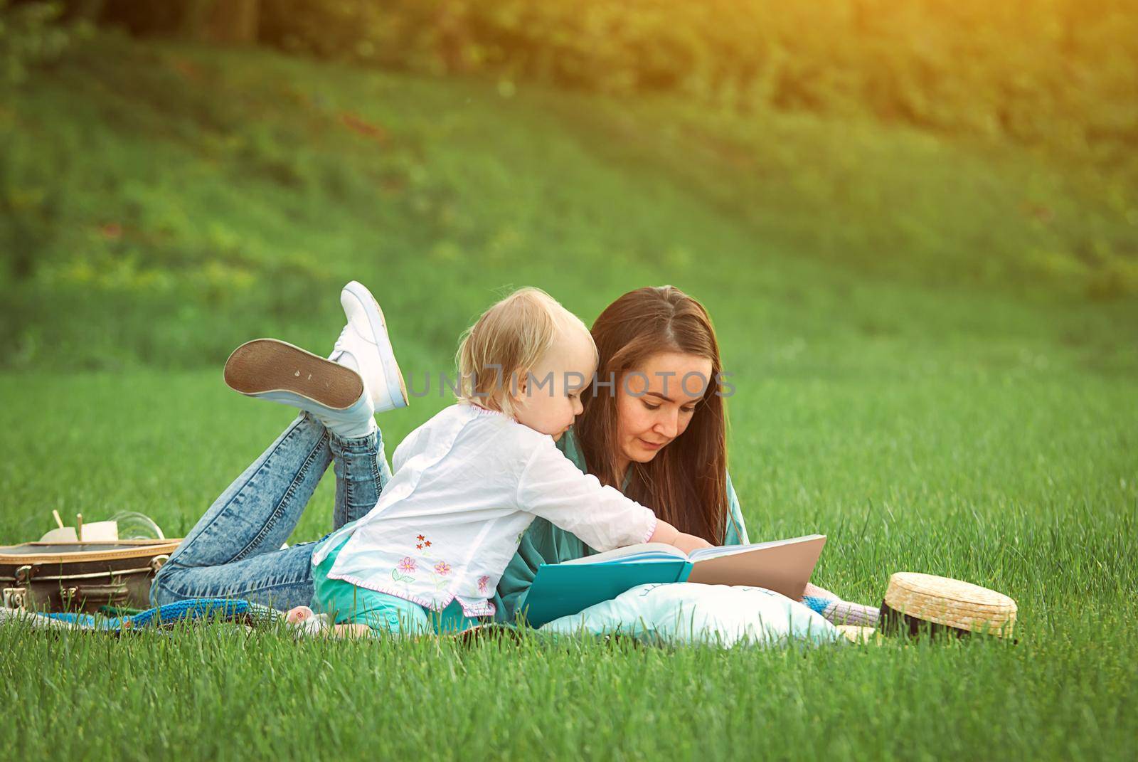 Mother is reading book for baby girl lying on the grass in the park by maramorosz