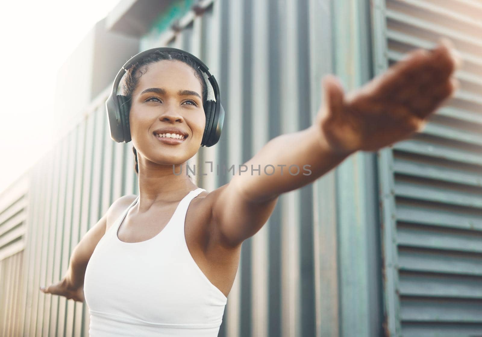 One fit young hispanic woman stretching arms in warrior pose for warmup to prevent injury while exercising in an urban setting outdoors. Happy and motivated female athlete listening to music with headphones while preparing body and mind for training workout or run by YuriArcurs