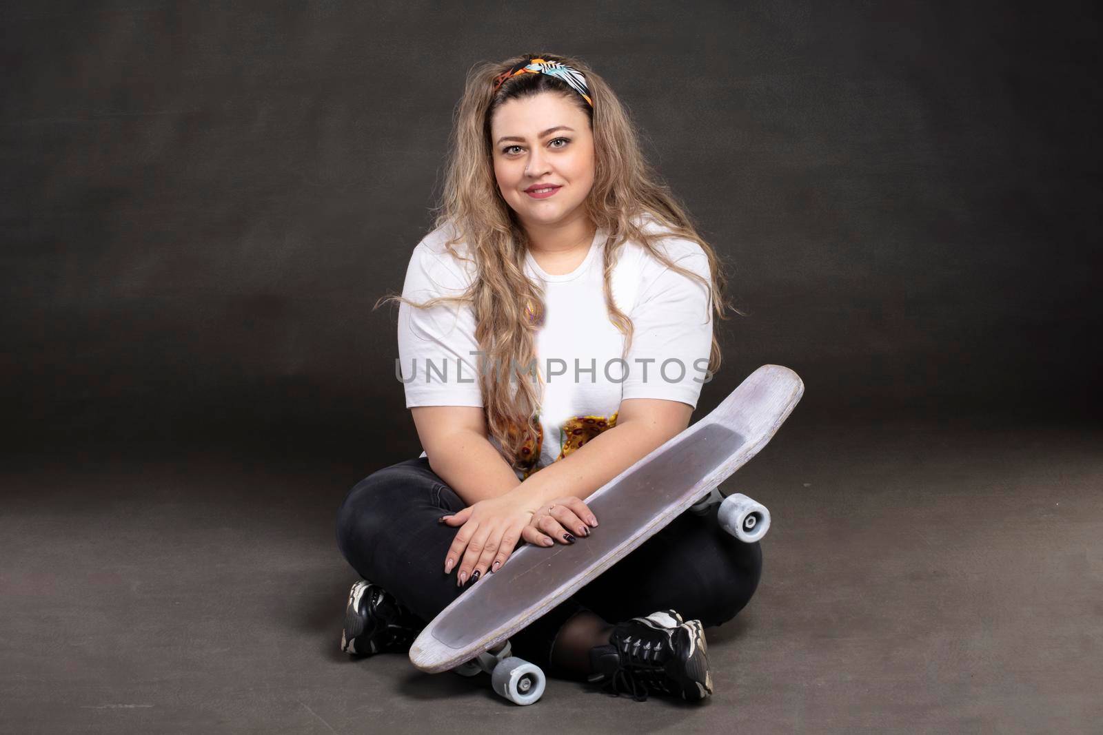 Beautiful fat woman with a skateboard on a gray background looks at the camera and smiles.