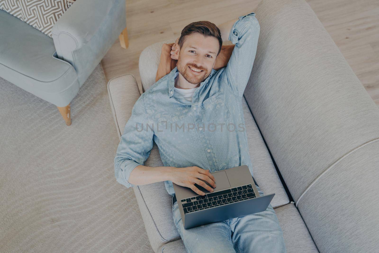 Happy young freelancer relaxing after finishing commission early, lying on comfortable couch in living room, resting head on hand, looking up while smiling and being satisfied