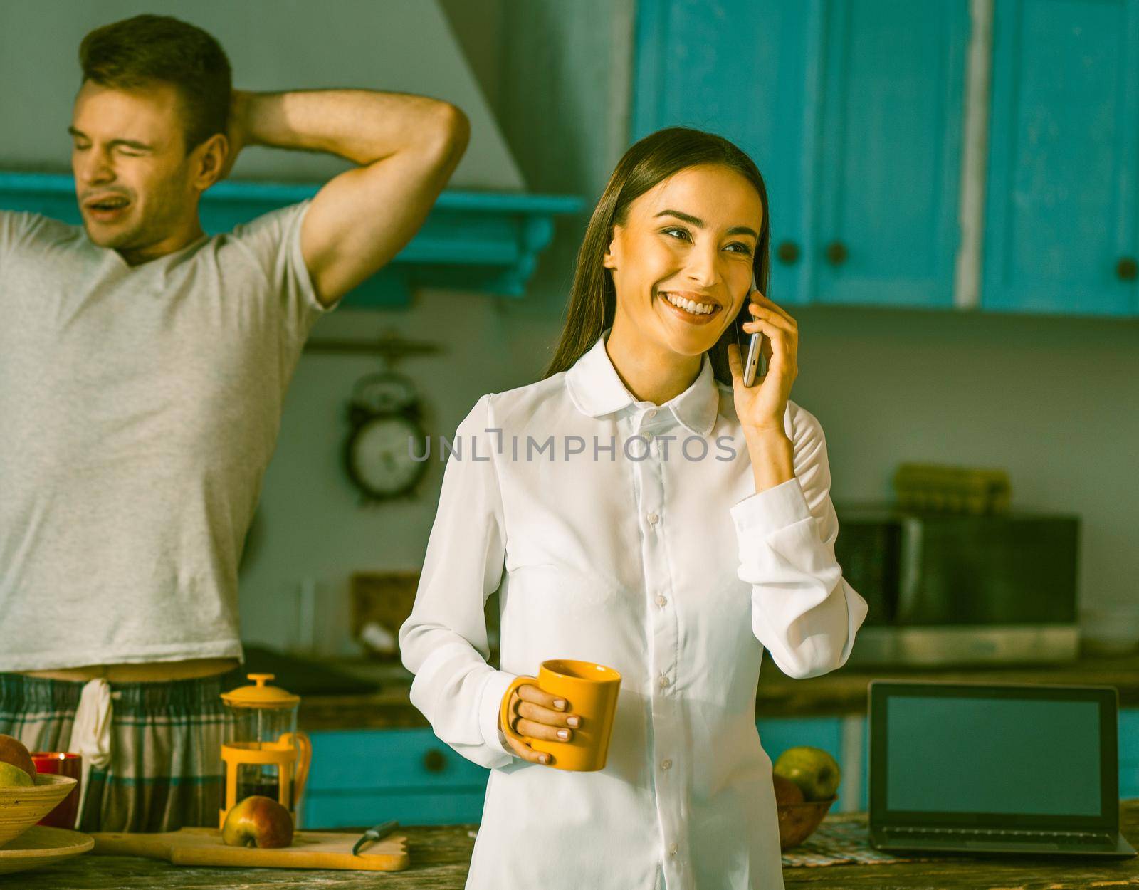 Happy couple chatting At Home kitchen In sunny morning, Smiling woman Talks Over Mobile Phone While Her Man In Pyjamas Yawns at background, Toned Image