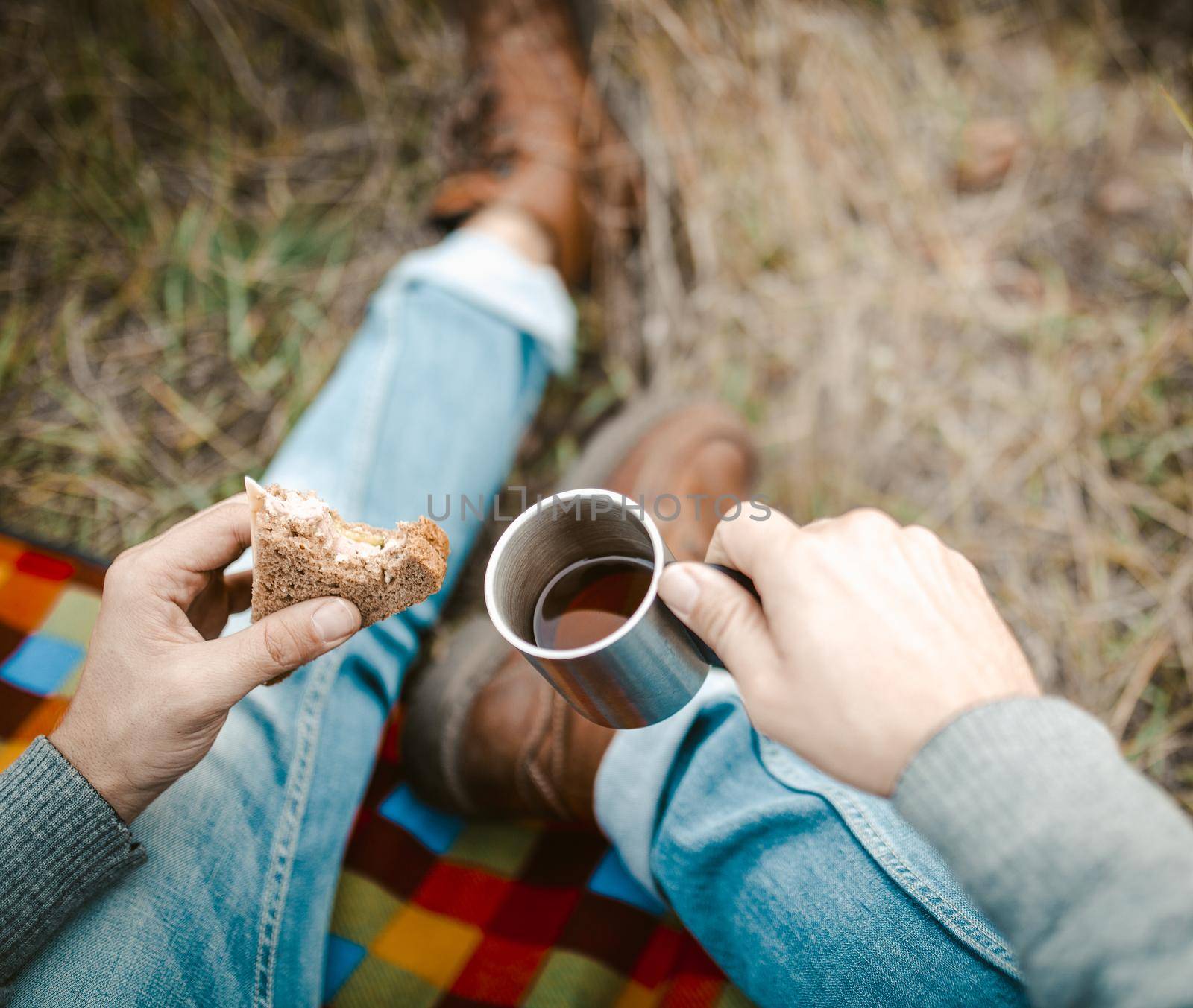 Young Man Eats Snack And Drinks Tea Sitting On Picnic Blanket At Grass, Focus On Caucasian Guy's Hands Holding Snack And Metal Cup With Hot Drink In Nature Outdoors, Close Up Shot, High Angle View