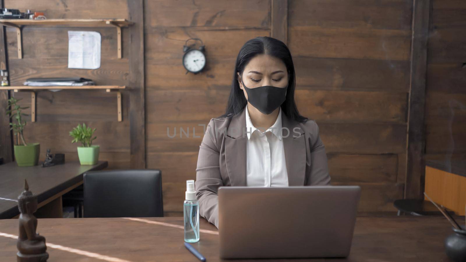 Lonely Business Woman Uses Computer Sitting At Table With Antiseptic Spray On It, Oriental Woman In Protective Mask Works With Laptop In Wooden Office Interior Isolated In Quarantine, Epidemic Concert