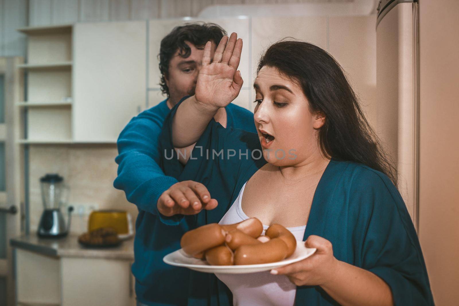 Wife Forbids Her Plus Size Husband To Take Sausage, Focus On Body Positive Brunette Woman Holding Plate Of Sausages In The Foreground, Her Man Reaches Out To Grab Sausage