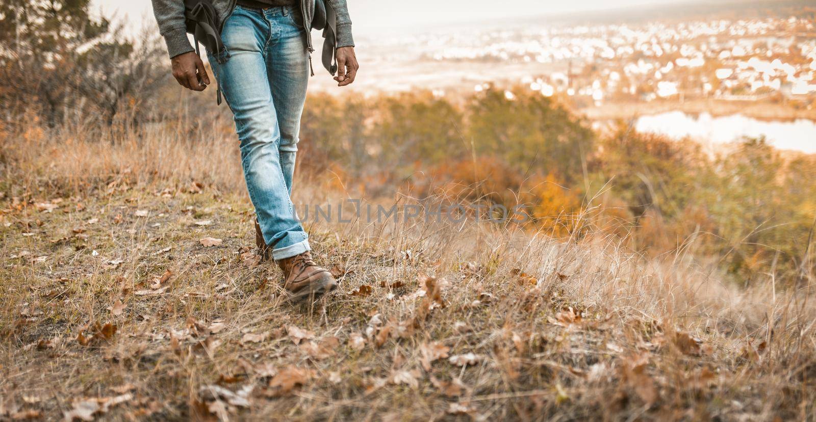 Man Hikes On A Hill, Young Man In Jeans With Backpack Is Walking Along The Autumn Grass With Beauty Landscape In Background, Close Up Of Men's Legs In Blue Jeans And Leather Boots.