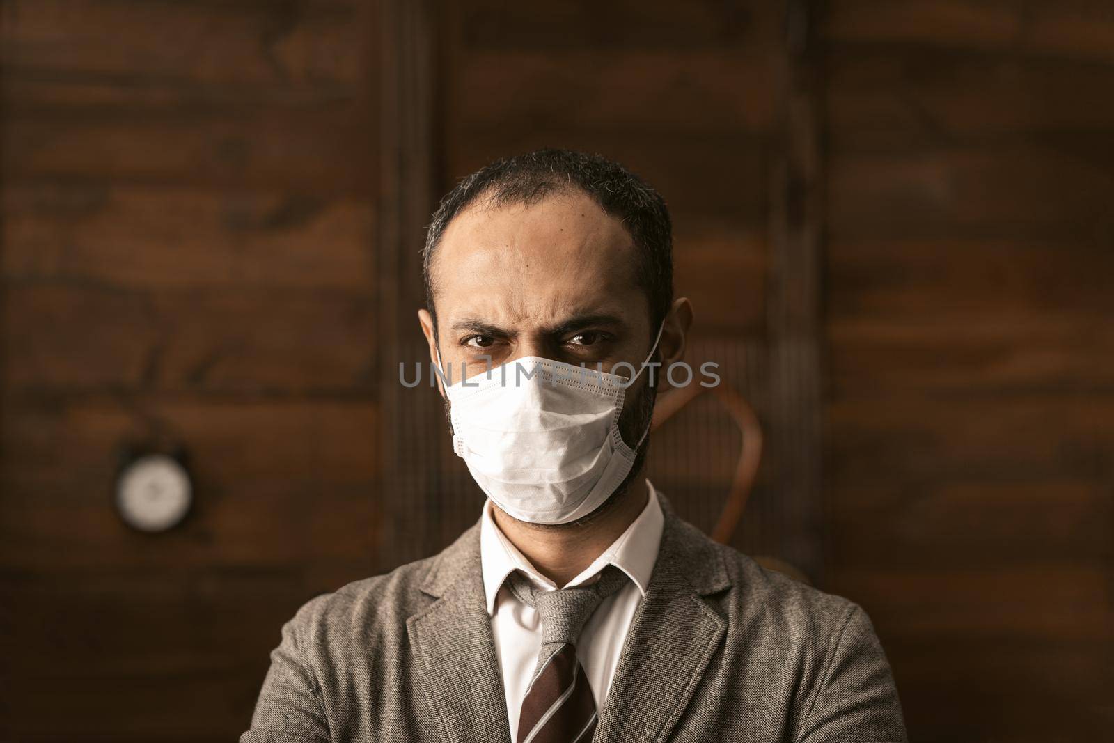 Serious Business Man In Protective Mask Is In Isolation Due To Epidemic Of Coronavirus, Middle Eastern Man In Suit Makes Stern Face Standing In Modern Office On Wooden Wall Backround, Toned Image