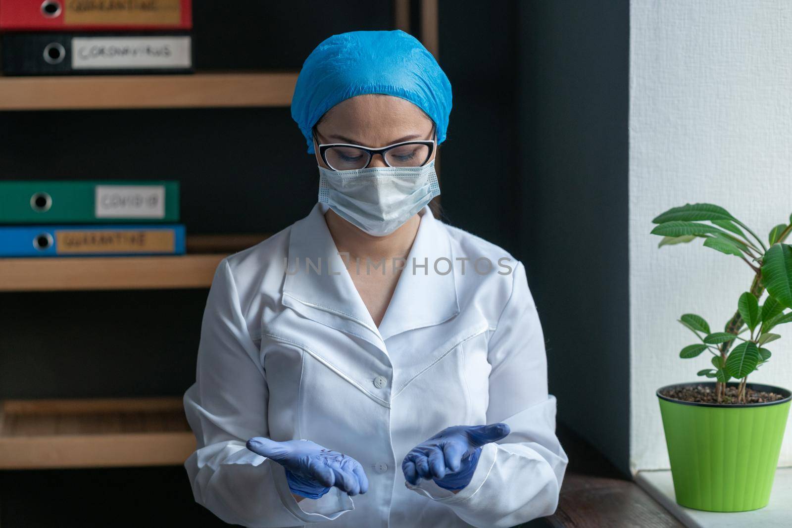 Woman In White Medical Uniform In Protective Mask And Gloves Carefully Researches Her Hands, Young Female Doctor Looks Down At Her Open Palms Through Glasses, Standing On Medical Office Back
