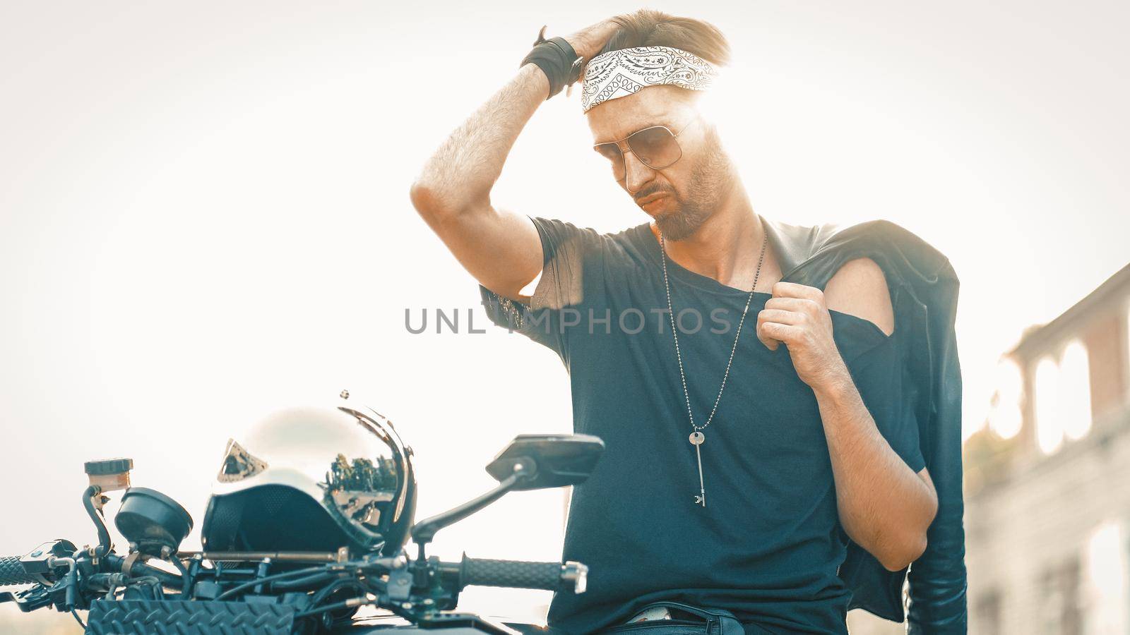 Stylish Biker In Sunglasses Is About To Ride Scooter From The Outside, Caucasian Man Holds Black Leather Jacket On His Shoulder While Standing In Backlight Near Motor Vehicle With Helmet On It