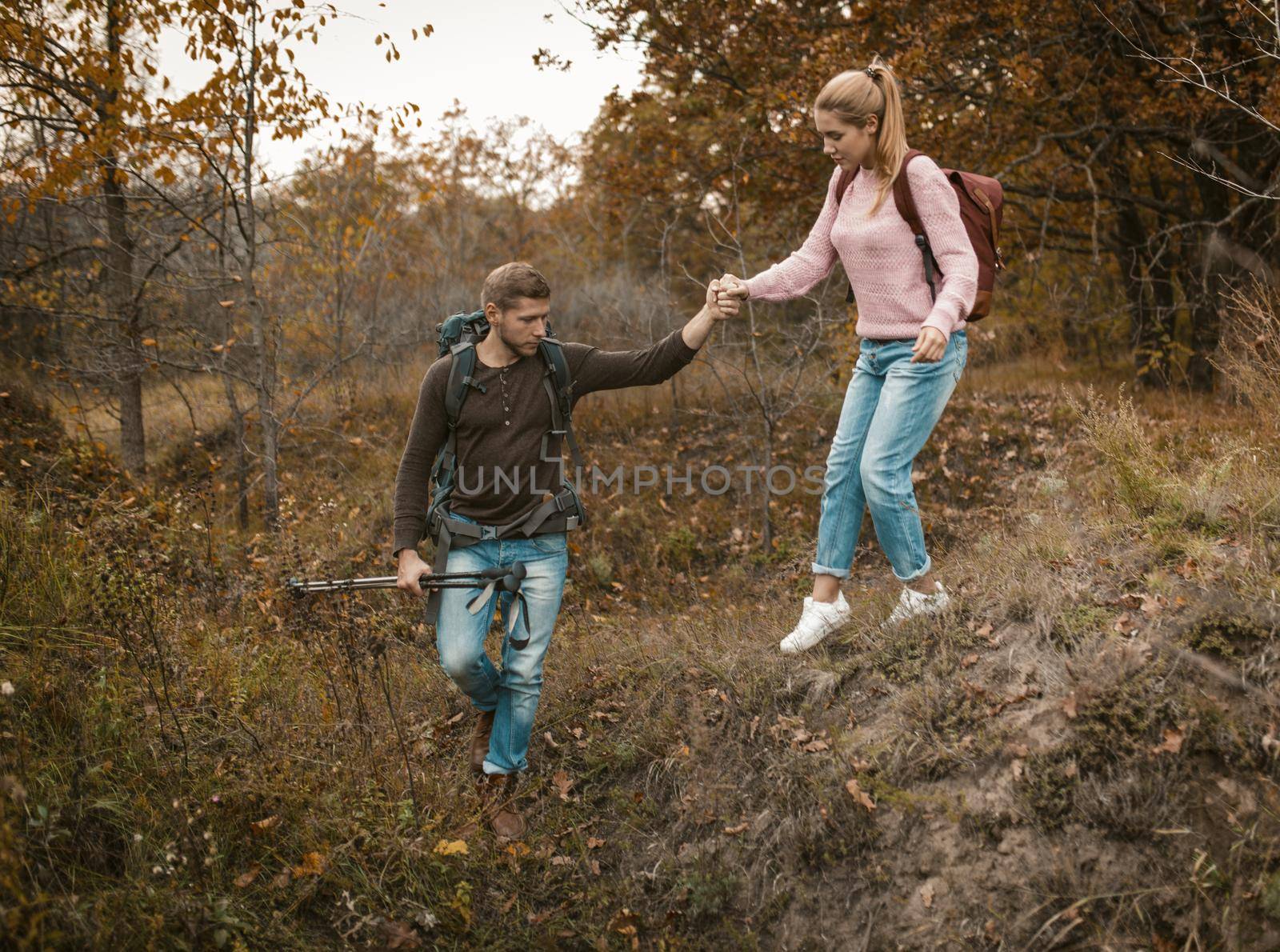 Young Couple Walks On Wooded Hill, Intelligent Caucasian Guy Supports With One Hand Of Girlfriend Go Down A Hill And Holds Hiking Poles With Other Hand