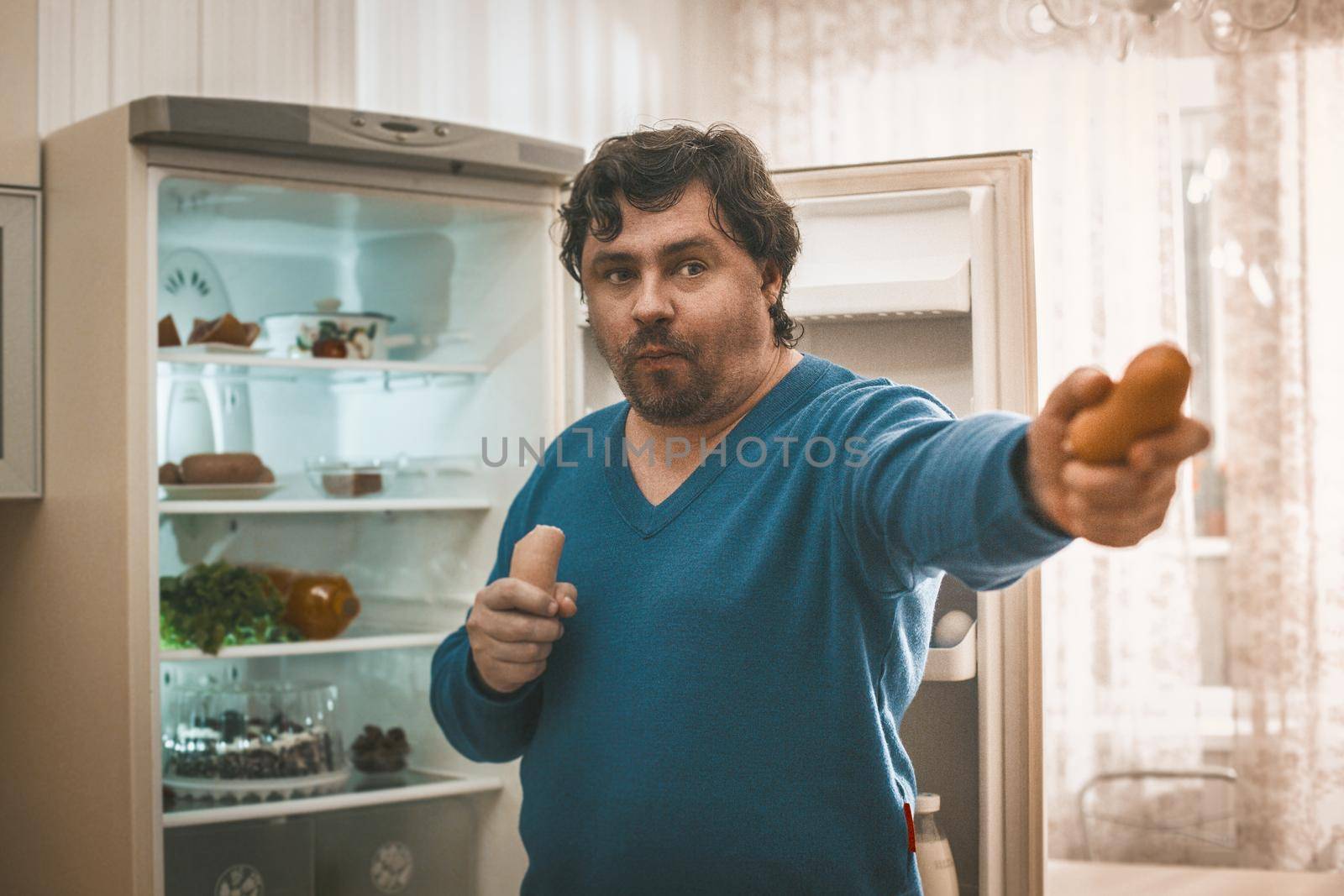 Plus Size Man Holds Out Sausage Into Camera, Selective Focused On Face Of Big Caucasian Man Eating And Holding Sausages In Hands While Standing Near Open Refrigerator At Home Kitchen Interior