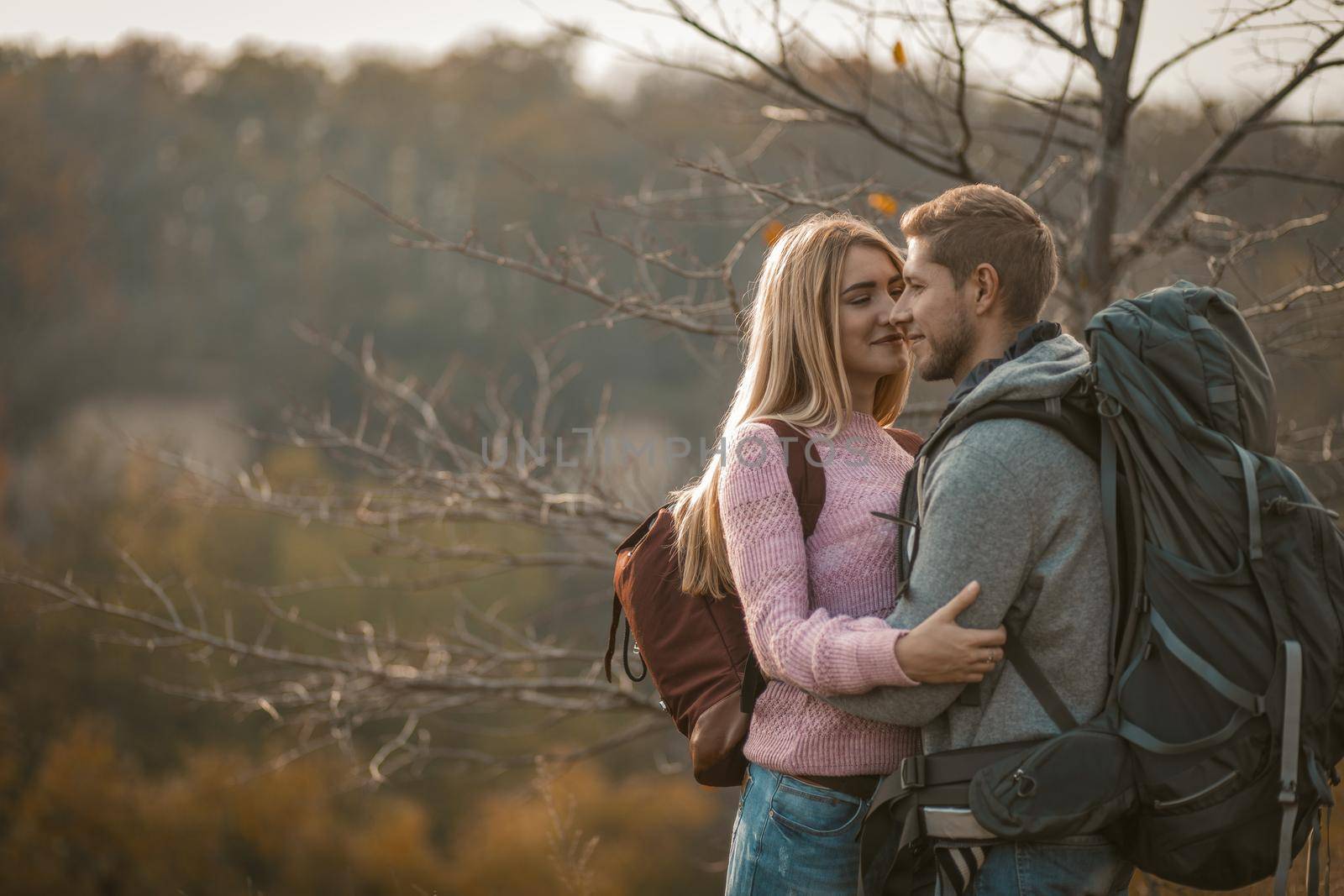 Happy Travelers With Backpacks Standing Embraced Against Background Of Autumn Nature, Young Caucasian Man And Woman Are Smiling While Looking At Each Other In Sunny Day, With Copy Space On Left Side