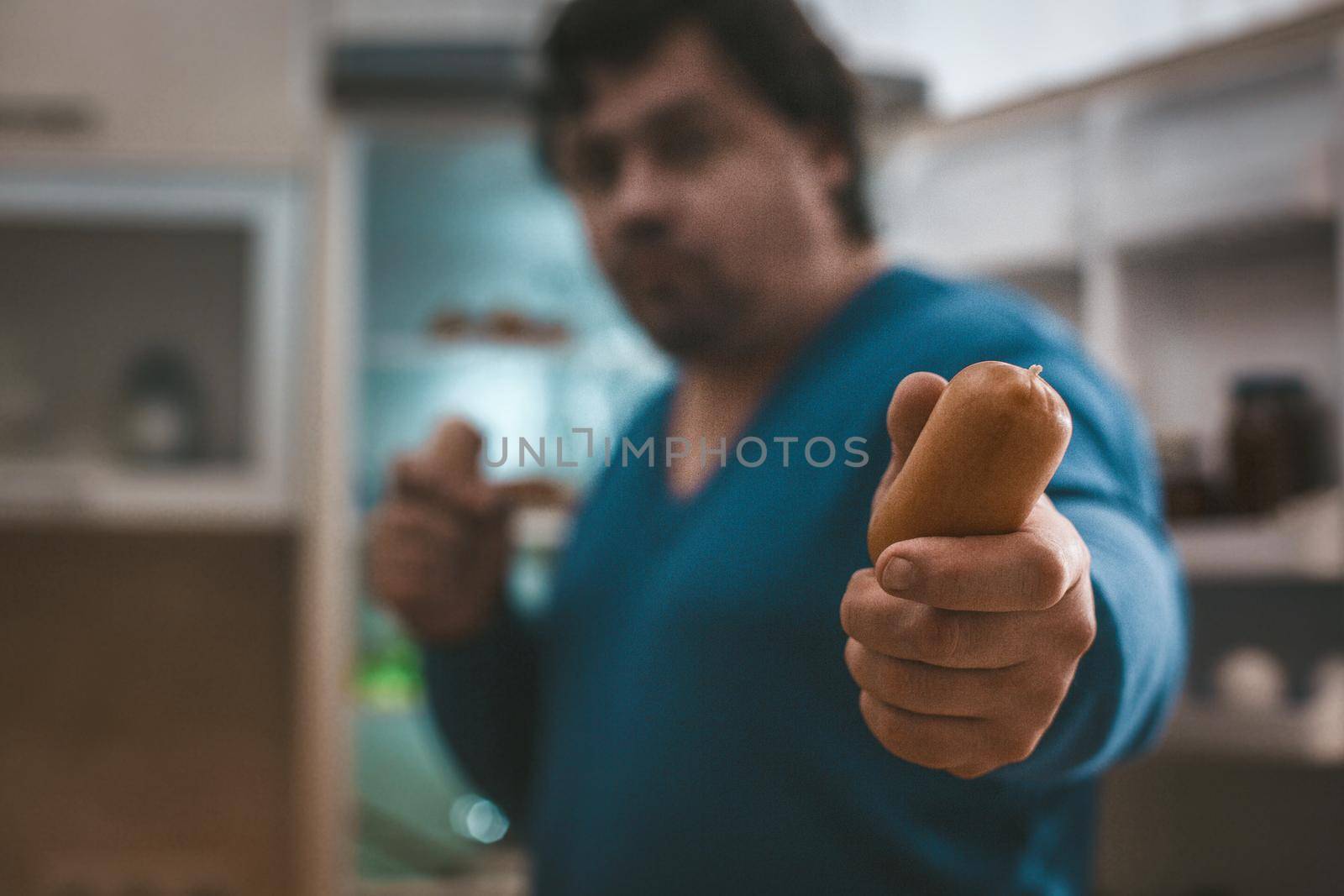 Plus Size Man Holds Out Sausage Into The Camera, Selective Focused On Sausage In Hand Of Big Caucasian Man In Blue Jumper Standing On Background Of Open Refrigerator, Close Up Shot