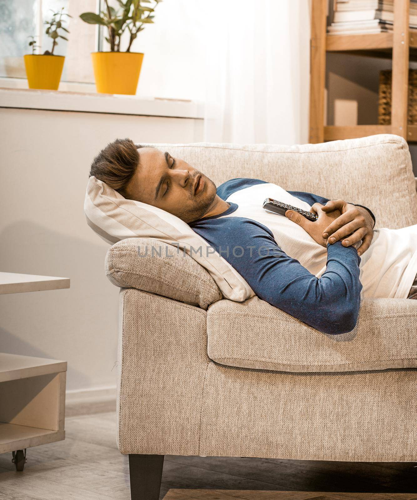 Man Relaxes On Couch At Home, Side View Of Sleeping Man In Home Wear Lying On Sofa Holding Remote Control With Hands, Young Man Staying At Home On Isolation During Quarantine, Toned Image