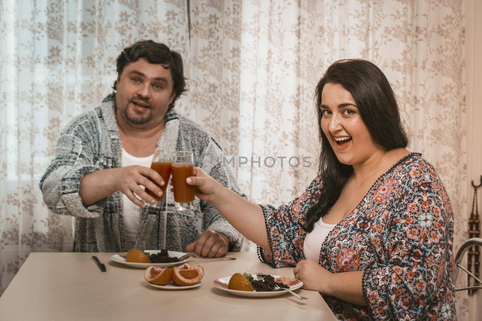 Body Positive Couple Clinking Glasses Of Juice While Sitting At Table With Wholesome Food, Plus Size Husband And Wife Cheerfully Looking At Camera While Sitting In Domestic Kitchen