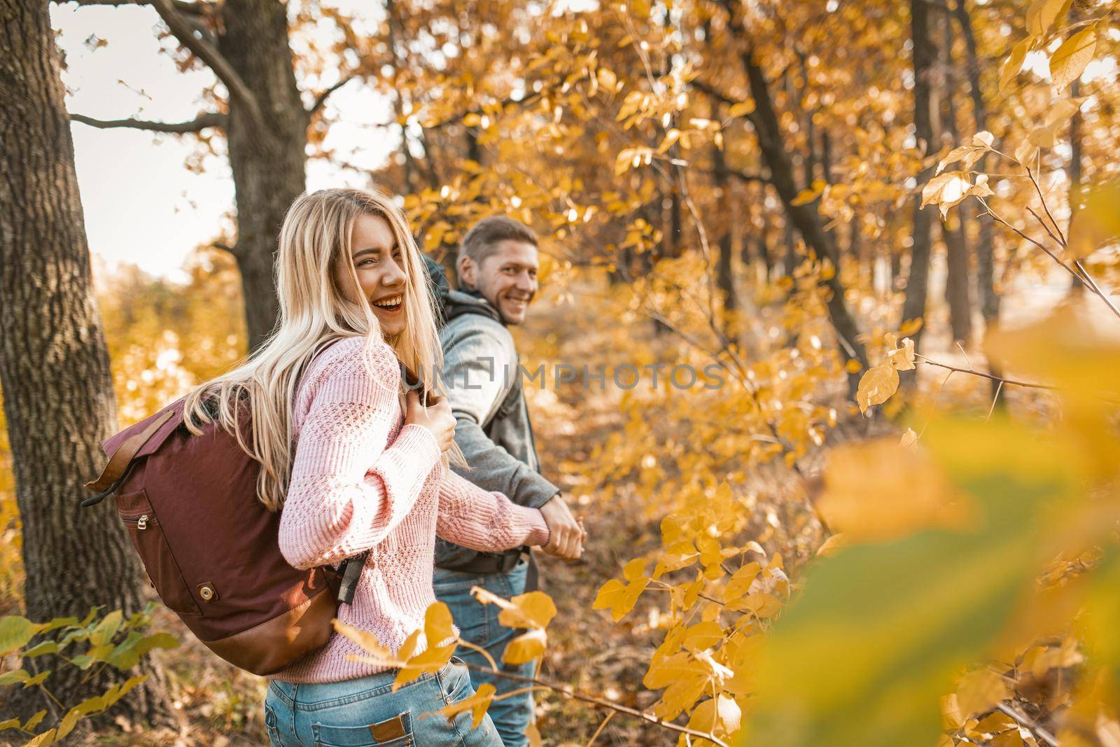 Happy Man And Woman Holding Hands Run Away Into Distance Along The Sunny Forest Path, Travelers Laughs Looking Back At Camera, Walking In Warm Sunny Day With Copy Space At Right Side
