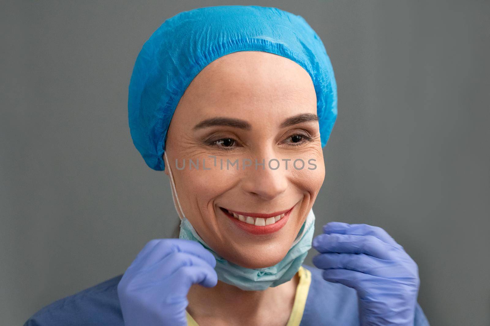 Smiling Woman In Medical Clothes Rejoices At End Of The Coronavirus Epidemic, Caucasian Woman In Blue Medical Uniform And Gloves Toothy Smiles While Taking Off Her Protective Mask, Quarantine Concept