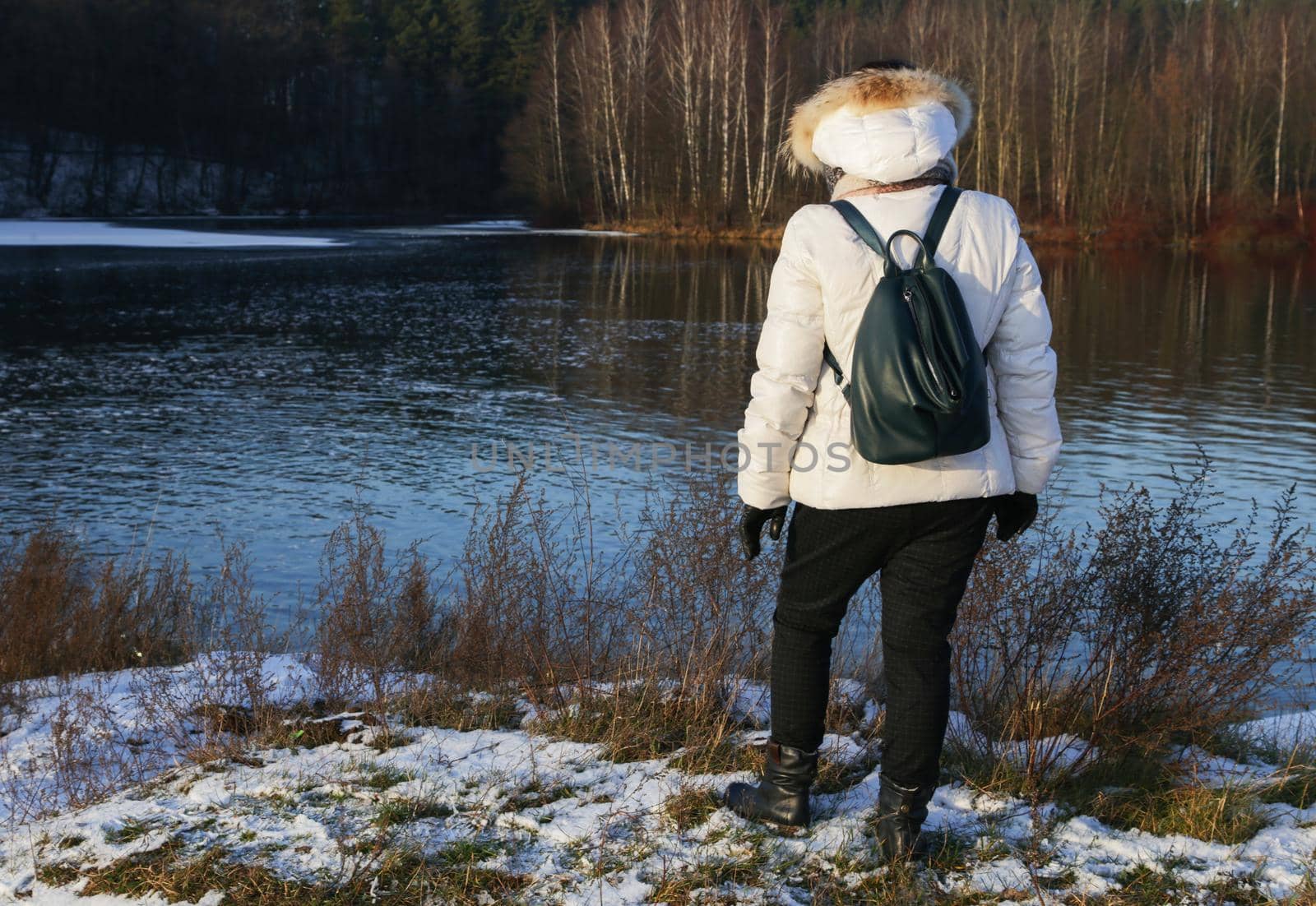 Woman in white jacket with dark backpack standing on the river bank with her back to the camera.