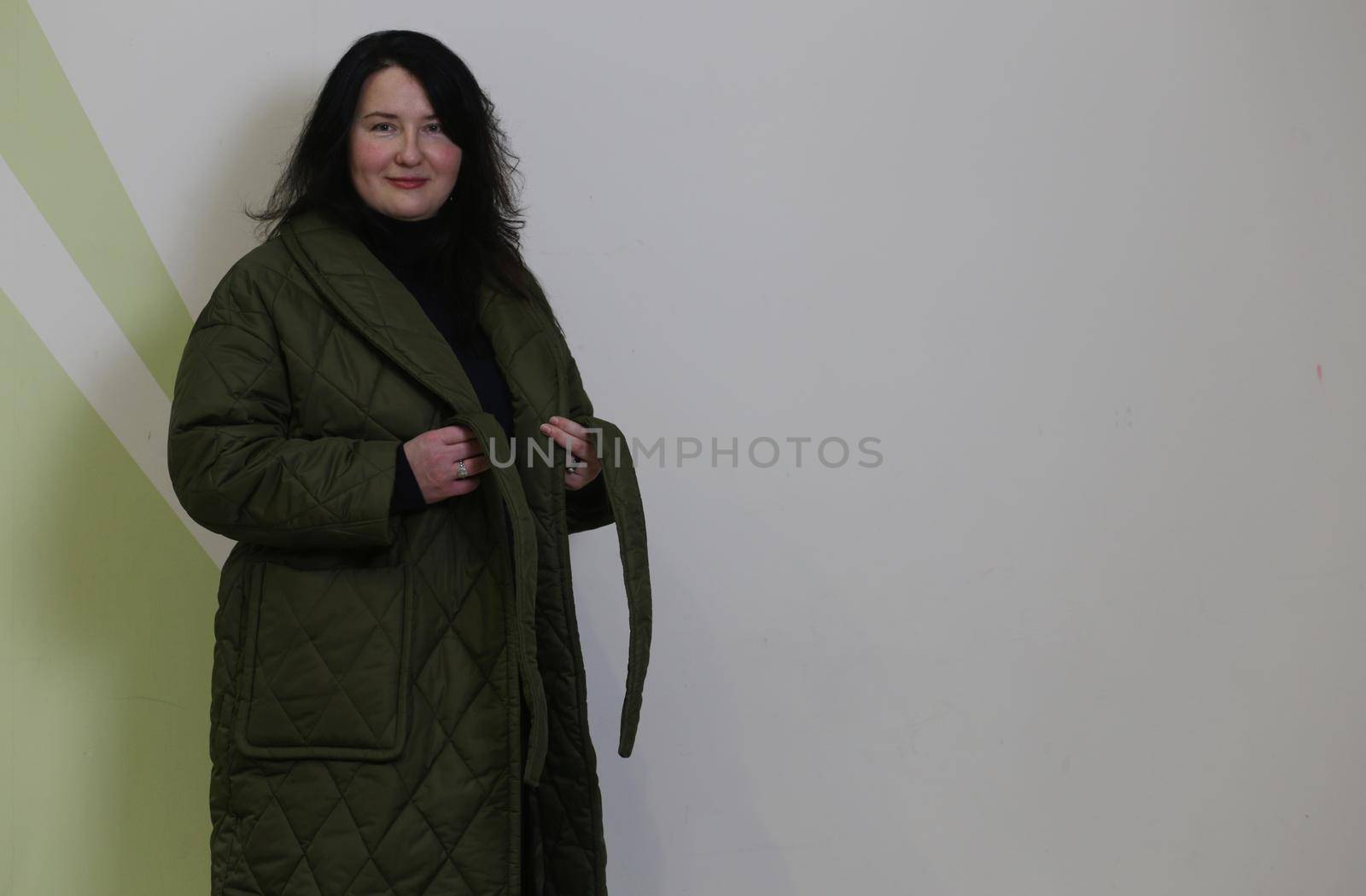 .A pretty plus size woman with black hair stands in a green raincoat against a white wall. A woman in an unbuttoned coat.