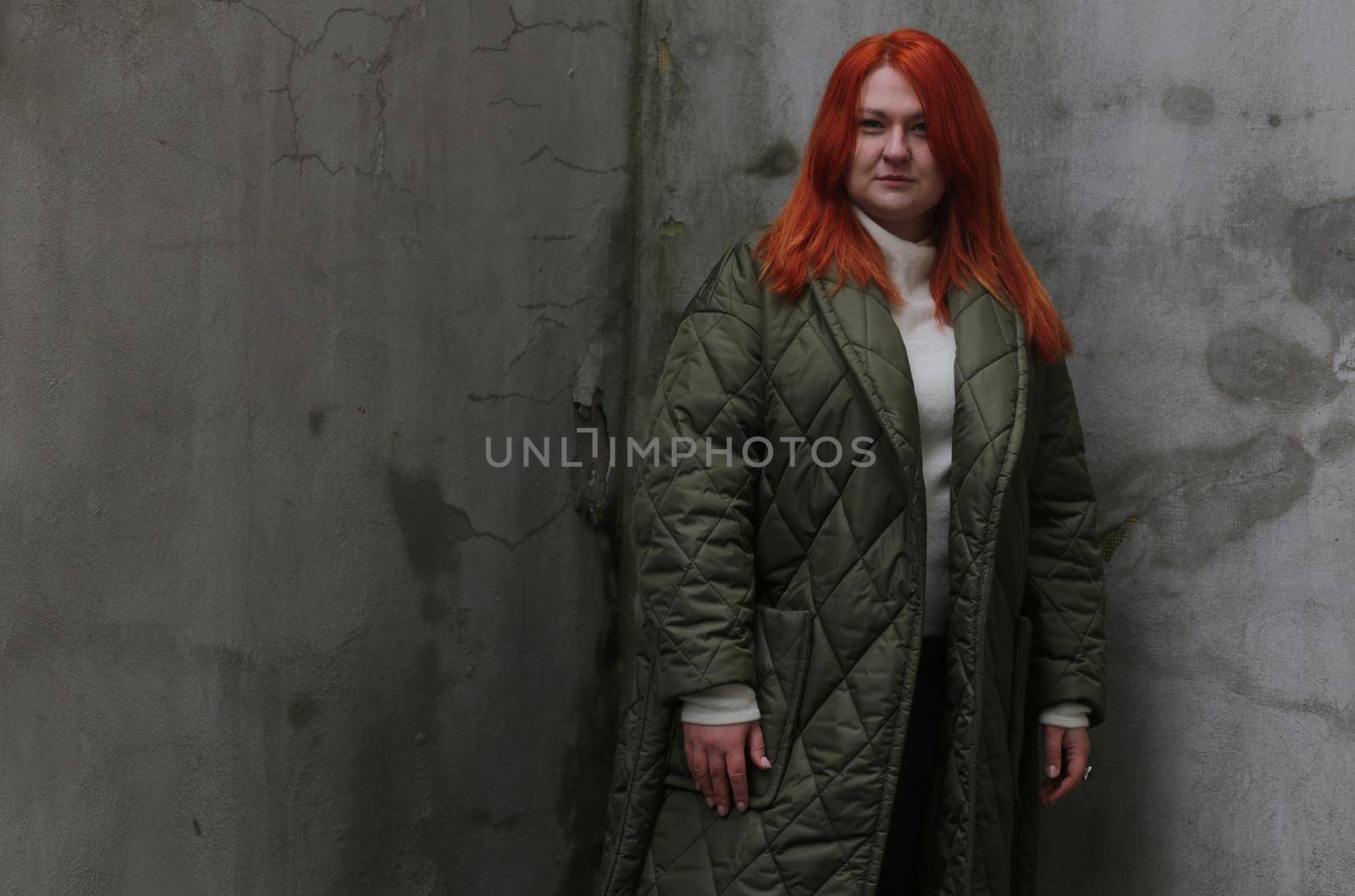 A plus-size girl with red hair in a green jacket poses in front of an old wall with damaged plaster.