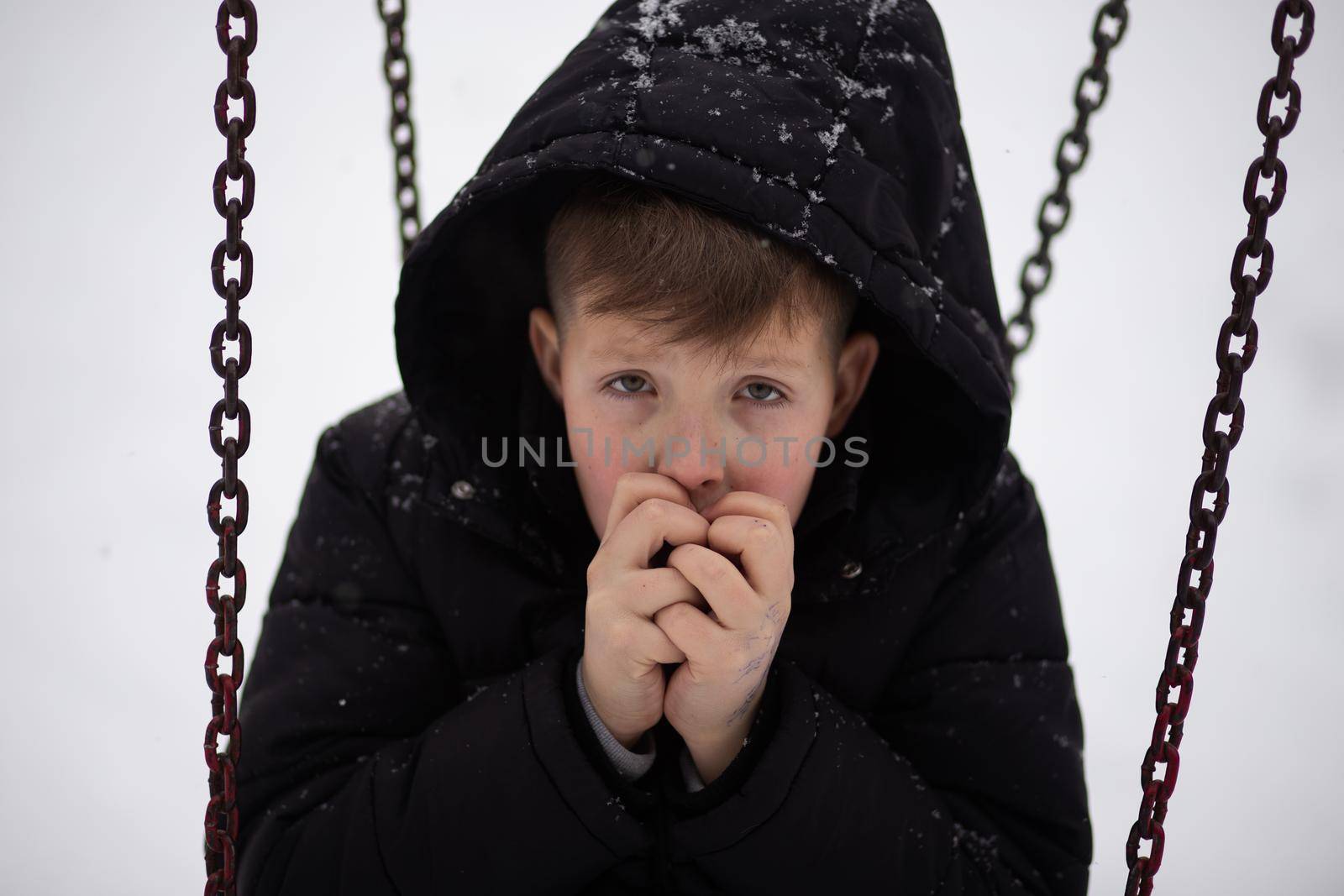 The boy in the hood holds his hands in front of his face. The boy warms his hands in winter. Freezing boy in an open space.