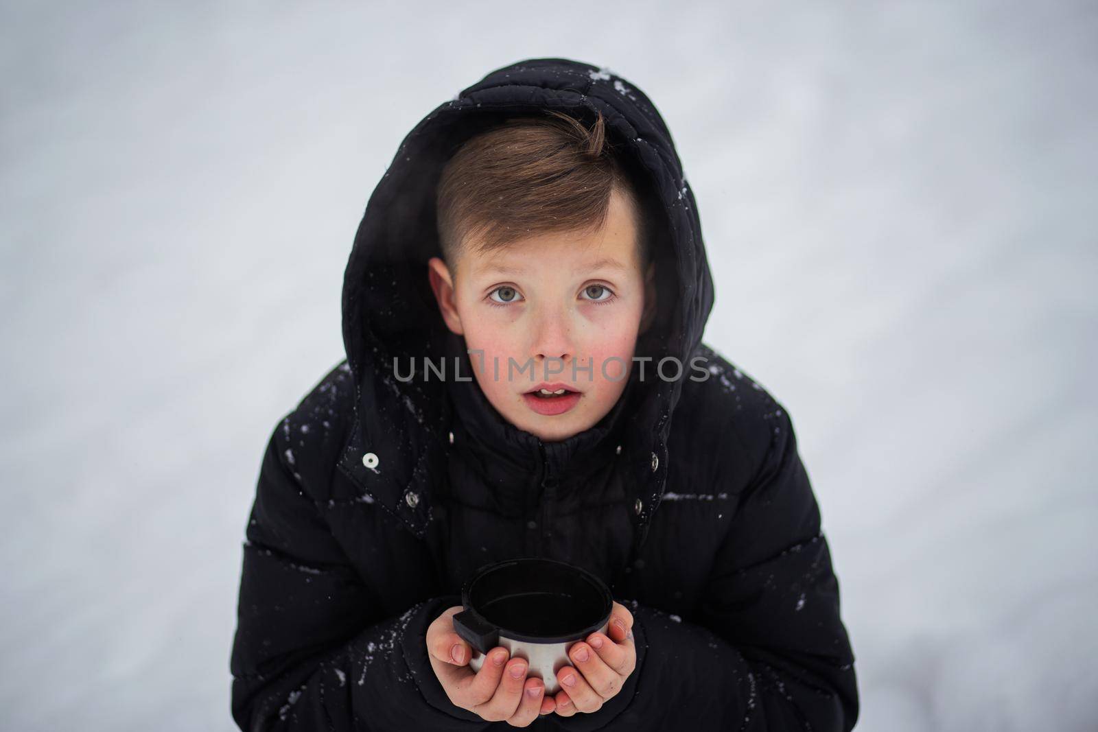 A cute teenage boy in winter clothes with a hood on his head is holding a metal mug in his hands. A handsome boy with a mug in his hands looks at the camera against a background of snow.