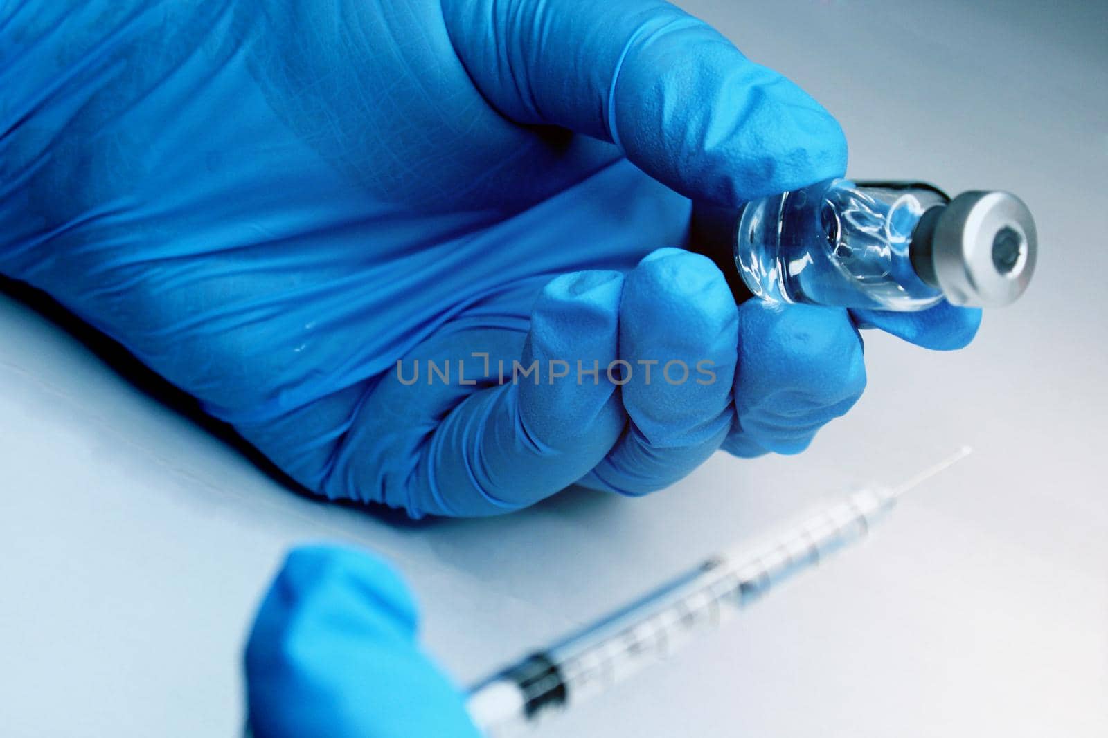 Close-up of a hand in a blue medical seal holding a vial of a vaccine. In the foreground, the syringe is out of focus. The concept of the fight against Covid19..