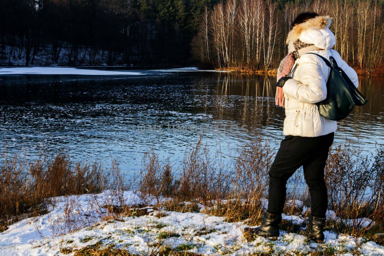 Side view of a woman in a white winter jacket against the background of the forest on the riverbank.