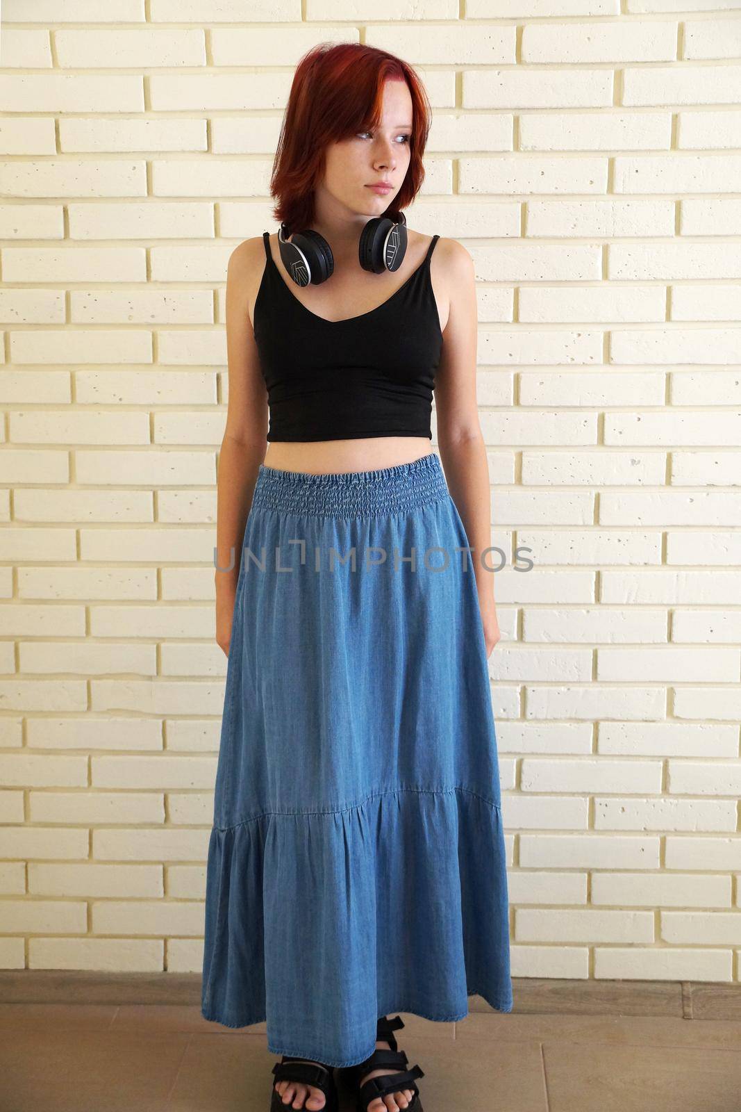 portrait of a red-haired teenage girl with headphones around her neck and in a blue long skirt against a brick wall.